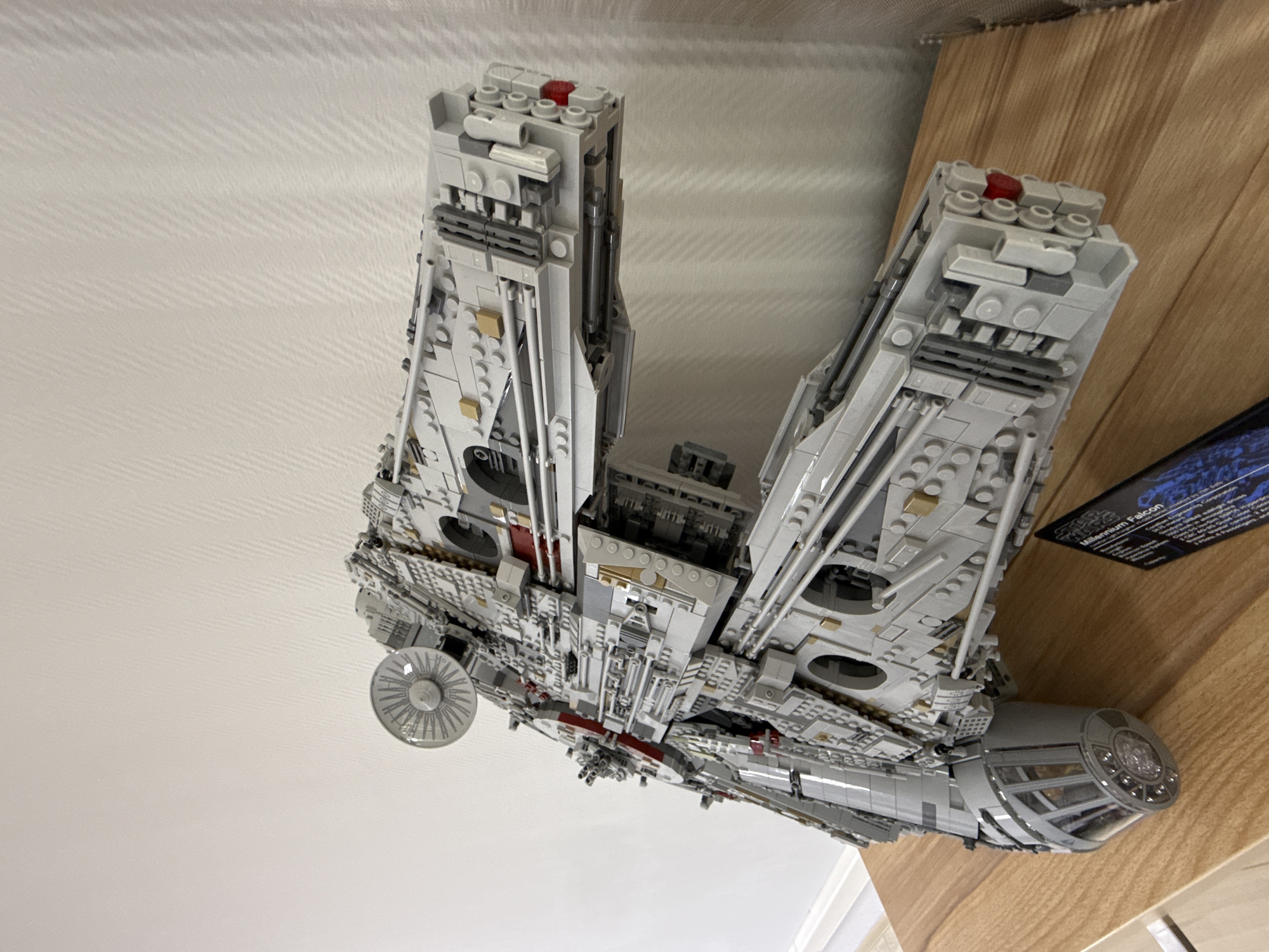 A high-angle shot depicts a Lego Millennium Falcon model resting partially on a wooden surface and partially against a white wall. The model is predominantly gray and composed of Lego bricks. The wooden surface is light brown and extends into the background. The white wall is smooth and featureless. No people are visible. The location is not identified as Krasnoyarsk, Russia.