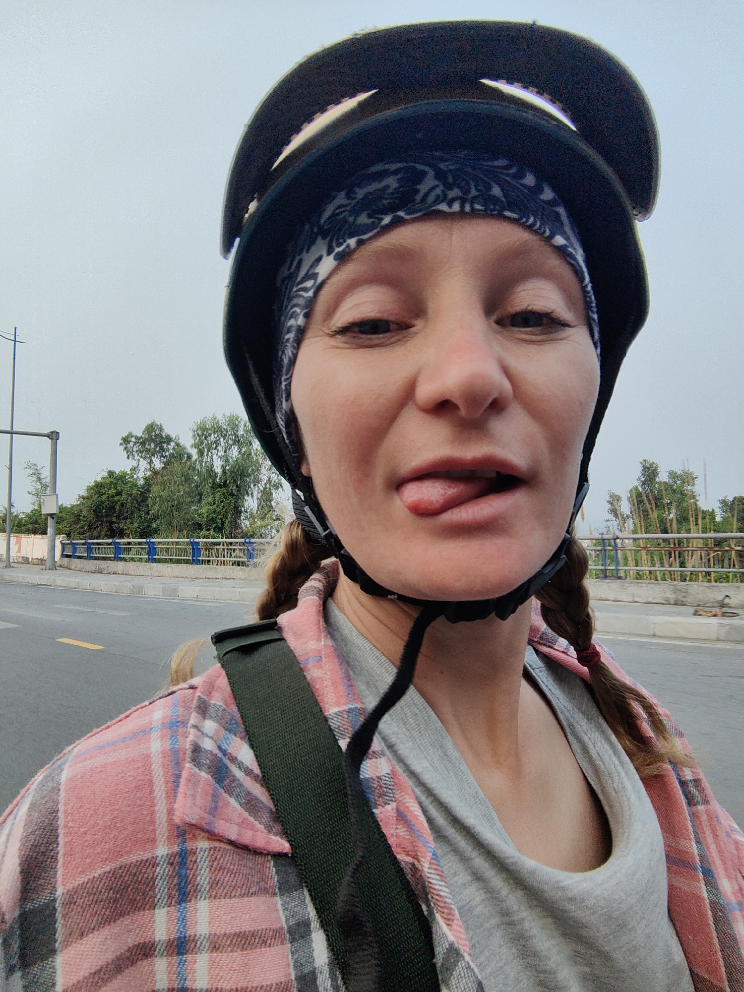 A medium, eye-level shot captures a fair-skinned individual with red hair pulled back in braids, wearing a black helmet over a patterned headscarf. The individual is sticking their tongue out. They are dressed in a grey v-neck shirt and a pink plaid flannel. A dark green strap crosses their shoulder.
The background suggests an outdoor urban setting in Bai Chay Ward, Vietnam, with a bridge, trees, a street with yellow markings, and a utility pole visible. The sky appears overcast.