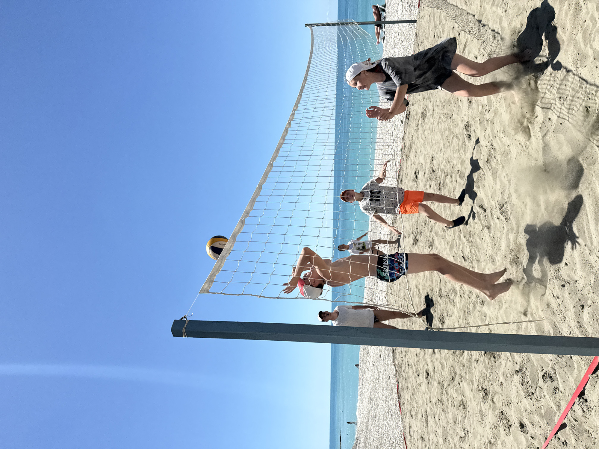 A group of young people are playing beach volleyball on a sunny day in Nebug, Russia. The volleyball net is set up on a sandy beach, with the turquoise Black Sea in the background. The sky is clear blue, and the sun is shining brightly. One player is in mid-air, attempting to hit the ball over the net, while others are positioned to receive it. The game is in full swing, with a sense of action and energy.