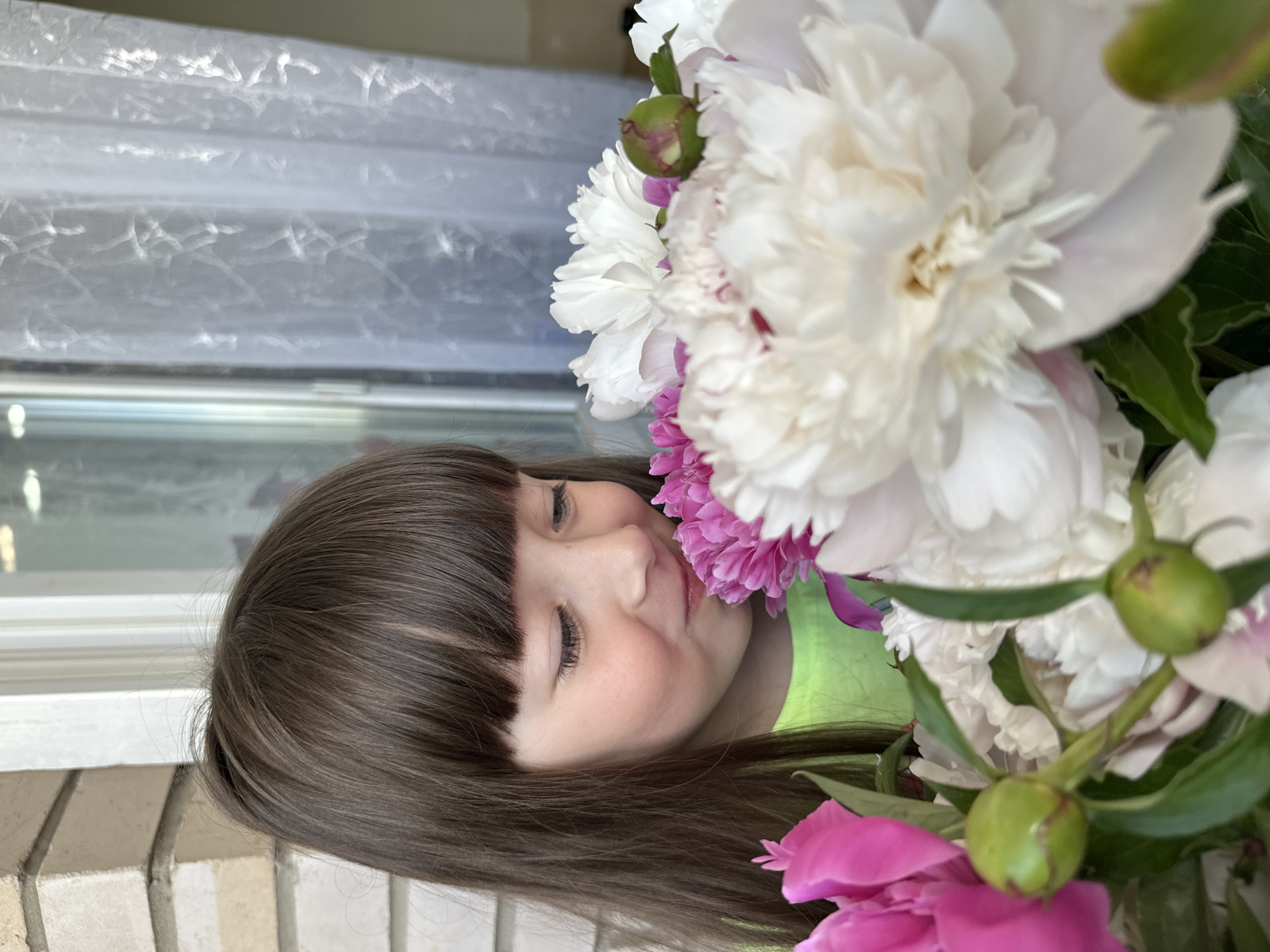A close-up, rotated portrait photograph shows a young girl with brown hair and bangs, eyes closed in a peaceful expression, nestled among large, showy peonies in shades of white and pink. The setting appears to be indoors, as evidenced by a window with a white, sheer curtain in the background, and a brick or stone wall visible on the left side of the frame. The location is Chaikovsky, Russia.