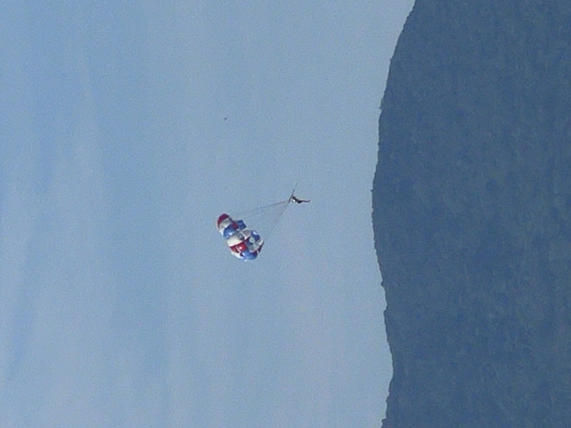The image shows a person parasailing high in the sky above a mountain range. The parasail is red, white, and blue. The sky is a clear blue. The mountain range is covered in green trees. The location is Đà Nẵng, Vietnam.