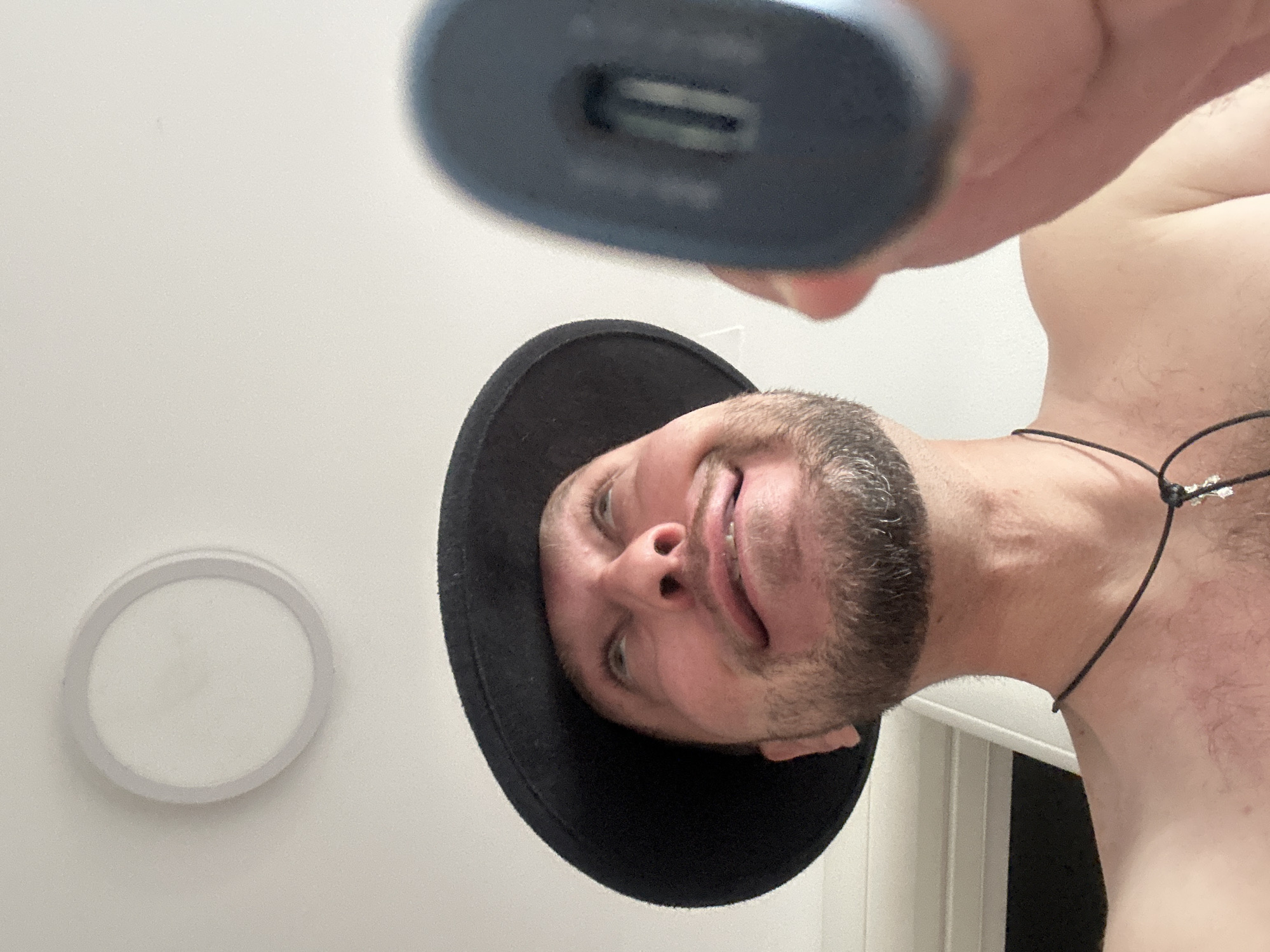 This media file captures a low-angle, upside-down selfie of a shirtless man wearing a black hat. The man has a short beard and appears to be smiling or smirking at the camera. He is holding a dark-colored electronic device, possibly a charger or power bank, which has a visible USB port. The background is a plain white wall, with a circular white light fixture visible on the left side of the frame. The lighting suggests an indoor setting, likely during the daytime. The overall mood is casual and playful, given the selfie's unusual perspective and the man's expression. There is no visible text in the image. The context provided places this scene in the Zvezdara Urban Municipality, Serbia.