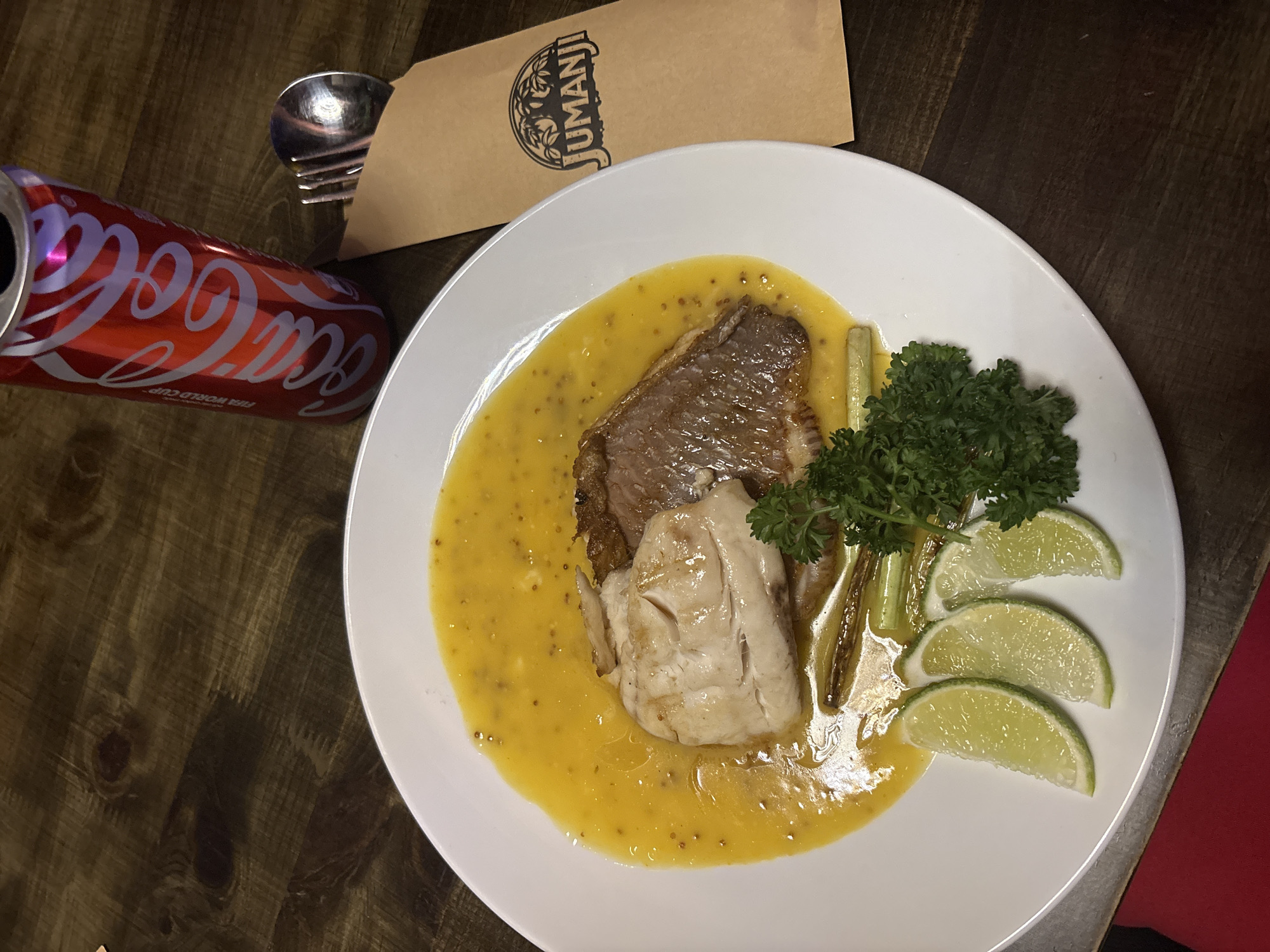The image shows a dish of food on a white plate, a Coca-Cola can, a fork and spoon, and a tan-colored menu or card with "Jumanji" printed on it, all placed on a dark wooden surface. The food on the plate appears to be a piece of cooked meat (likely beef) layered over a piece of cooked fish, surrounded by a yellow sauce with mustard seeds, garnished with parsley and lime wedges. The Coca-Cola can is open and partially obscured. The fork and spoon are lying next to the menu. The setting is most likely a restaurant in Nha Trang Ward, Vietnam.