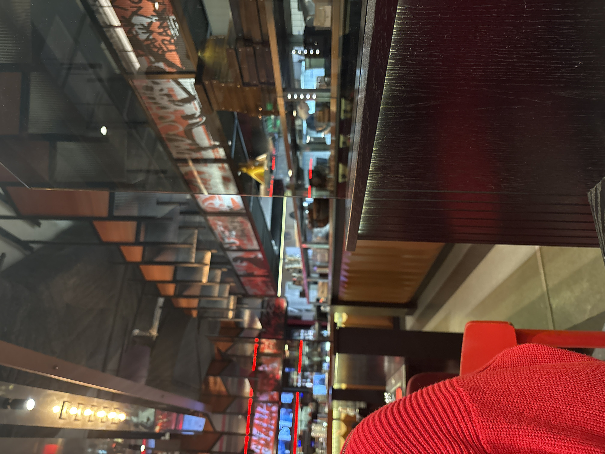 The image captures a view within a multi-level establishment, possibly a restaurant or bar, in Saint Petersburg, Russia. The foreground is dominated by a dark wooden surface, likely a countertop or tabletop. In the lower right, a bright red knitted garment, possibly a sweater or scarf, is visible, suggesting a person is present at the scene.
Through a glass barrier or reflection, a complex interior is revealed. The layout includes multiple levels connected by stairs. Red vertical lines, possibly decorative lighting or accents, run through the interior, adding to the visual complexity. There are posters and advertisements, and there are industrial elements like bare metal beams. The building appears to be well lit with warm lighting that reflects off the surfaces creating some light distortion. In the upper middle of the image, the blurred figure of at least one individual is seen, adding a human presence to the scene.