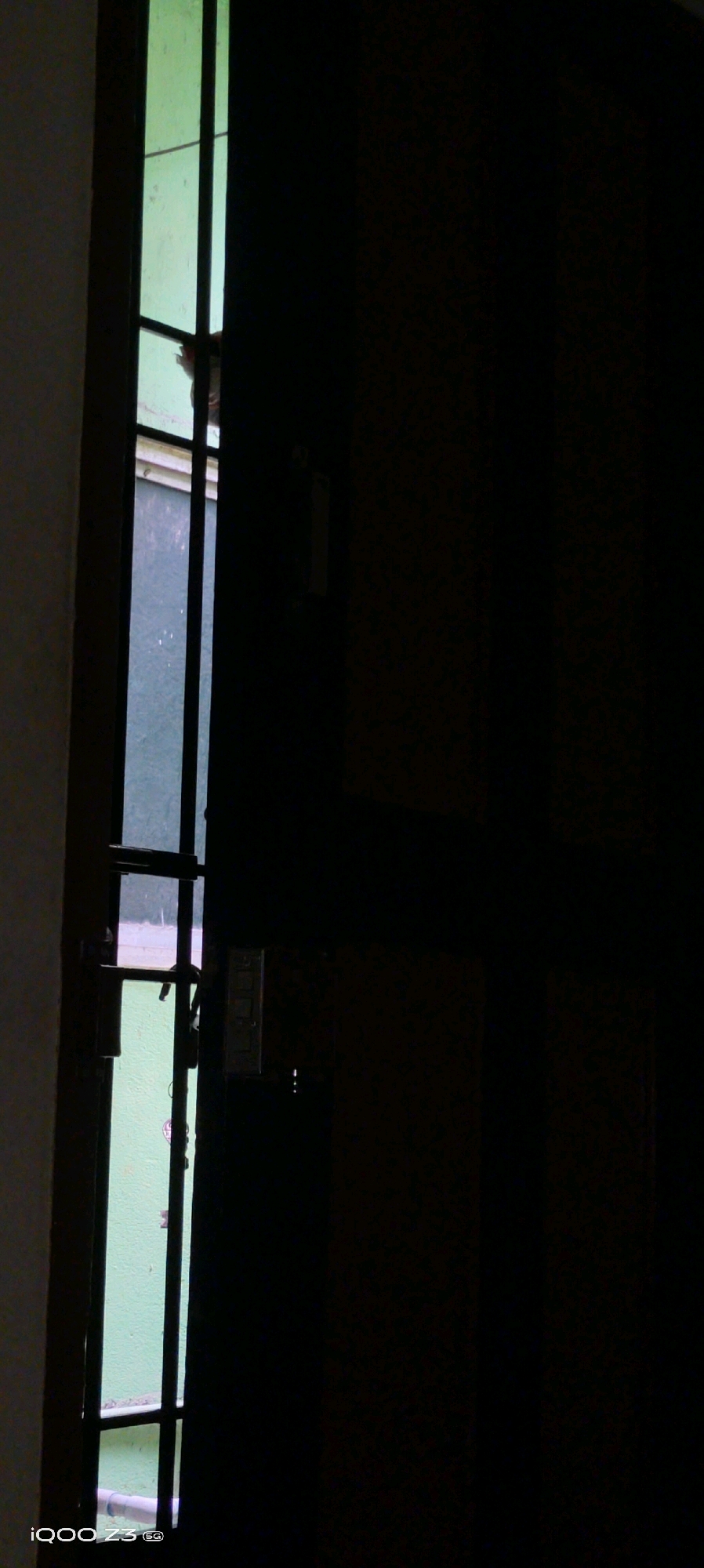 The image depicts a partially opened dark wooden door revealing a sliver of a light green wall and vertical bars, possibly from a window or gate. The lighting suggests an indoor setting, with the right side of the frame dominated by the dark door, and the left side showing the exterior or another space through the bars. A portion of a light-colored object, possibly a pipe or tube, is visible at the bottom left. The bottom left corner of the image contains a watermark that reads "iQOO Z3 5G". The scene is devoid of any people or discernible activity. The specific location in Chennai, India, cannot be determined from the visual information presented.