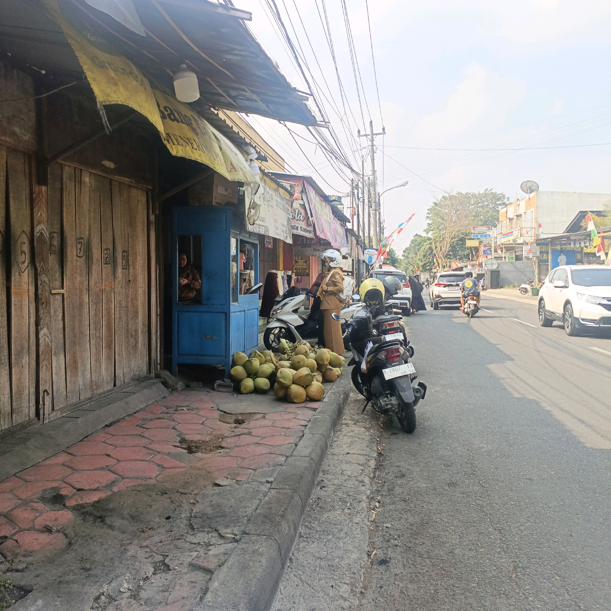 The image depicts a street scene in Salatiga, Indonesia, likely during daylight. On the left, a weathered wooden building with numbers painted on its doors stands adjacent to a blue stall. A person, partially visible through the stall's window, appears to be attending to it. In front of the stall, a pile of coconuts is arranged on the patterned red and grey sidewalk. Two motorcycles are parked on the side of the road, one white and one black, with helmets placed on them. A woman in a brown uniform and white helmet stands next to the white motorcycle, facing the street. Beyond the stall, other businesses with awnings and signage are visible, with flags of Indonesia flying. Cars and motorcycles are present on the asphalt road, indicating traffic. Power lines crisscross the sky, supported by utility poles. The overall environment suggests a commercial area within the city, with elements of daily life and commerce.