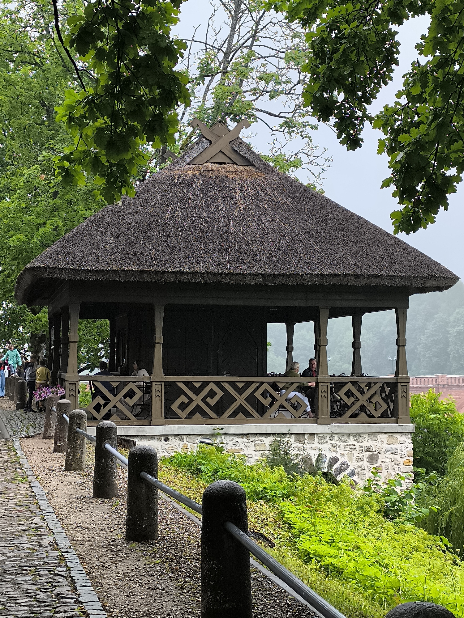 The image depicts a charming thatched-roof gazebo situated in the historic town of Kuldīga, Latvia. The gazebo features intricate wooden lattice railings and is supported by sturdy wooden posts. Inside, a couple is seated at a table, engaged in conversation, while other visitors are visible walking along a cobblestone path to the left of the structure. The gazebo is perched on a stone foundation, surrounded by lush green foliage. The overall atmosphere is tranquil and inviting, suggesting a peaceful day in this picturesque European town.