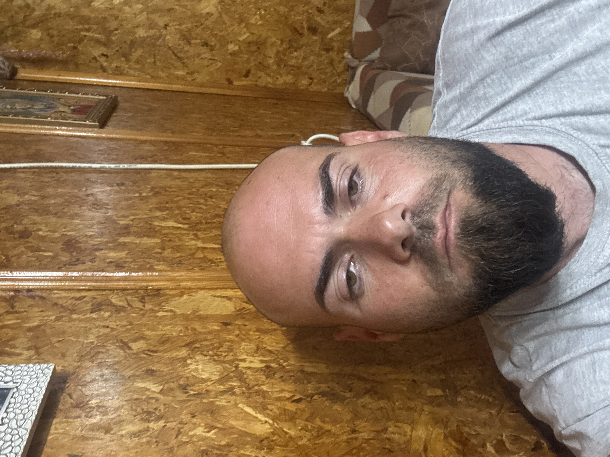 A close-up, rotated image shows a bald, bearded man wearing a gray t-shirt. The background appears to be a wall made of wood. A white cable runs along the wall. Two framed images are visible in the background. The location in the city of Sarnevo, Bulgaria, cannot be determined from the visual data.