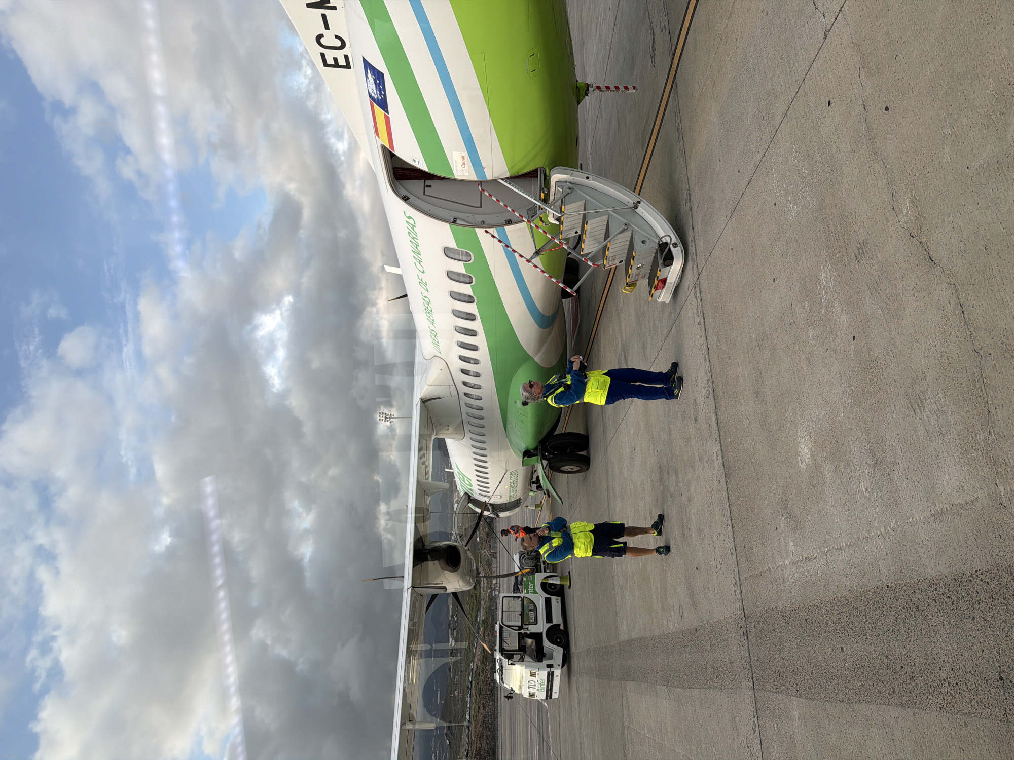 The image captures an active airport tarmac scene in Granadilla de Abona, Spain, during daytime under a partly cloudy sky. The central subject is a turboprop aircraft, possibly a regional airliner, with a distinctive livery featuring green, white, and light blue stripes.
The aircraft's main cabin door is open, revealing integrated airstairs extending to the ground. Red and white striped cones, bearing the text "REMOVE BEFORE FLIGHT," are placed around the bottom of the stairs. Visible text on the fuselage includes "EC-M" near the tail, indicating its registration, and "LINEAS AEREAS DE CANARIAS" along the side. A flag resembling the Spanish national flag with a coat of arms is also seen on the tail section. Inside the open door, the word "Canair" is visible.
Two ground crew members are prominent in the foreground. Both wear high-visibility yellow vests over dark blue attire, with one in shorts and the other in long pants. The crew member closer to the front of the plane stands near a white pushback tug, which has "Better" written on its side. This individual holds what appears to be a marshalling wand or headset. The second crew member stands further back, near the aircraft's engine and propeller, also holding a similar item. A third individual in an orange vest is partially visible behind the second crew member.
The ground is paved concrete or asphalt, showing various markings and textures. In the background, beyond the aircraft's wing, distant mountains are visible under the cloudy sky, possibly reflected in the wing's surface or seen through a window. The scene suggests activities related to aircraft ground operations, such as preparing for departure or servicing upon arrival.