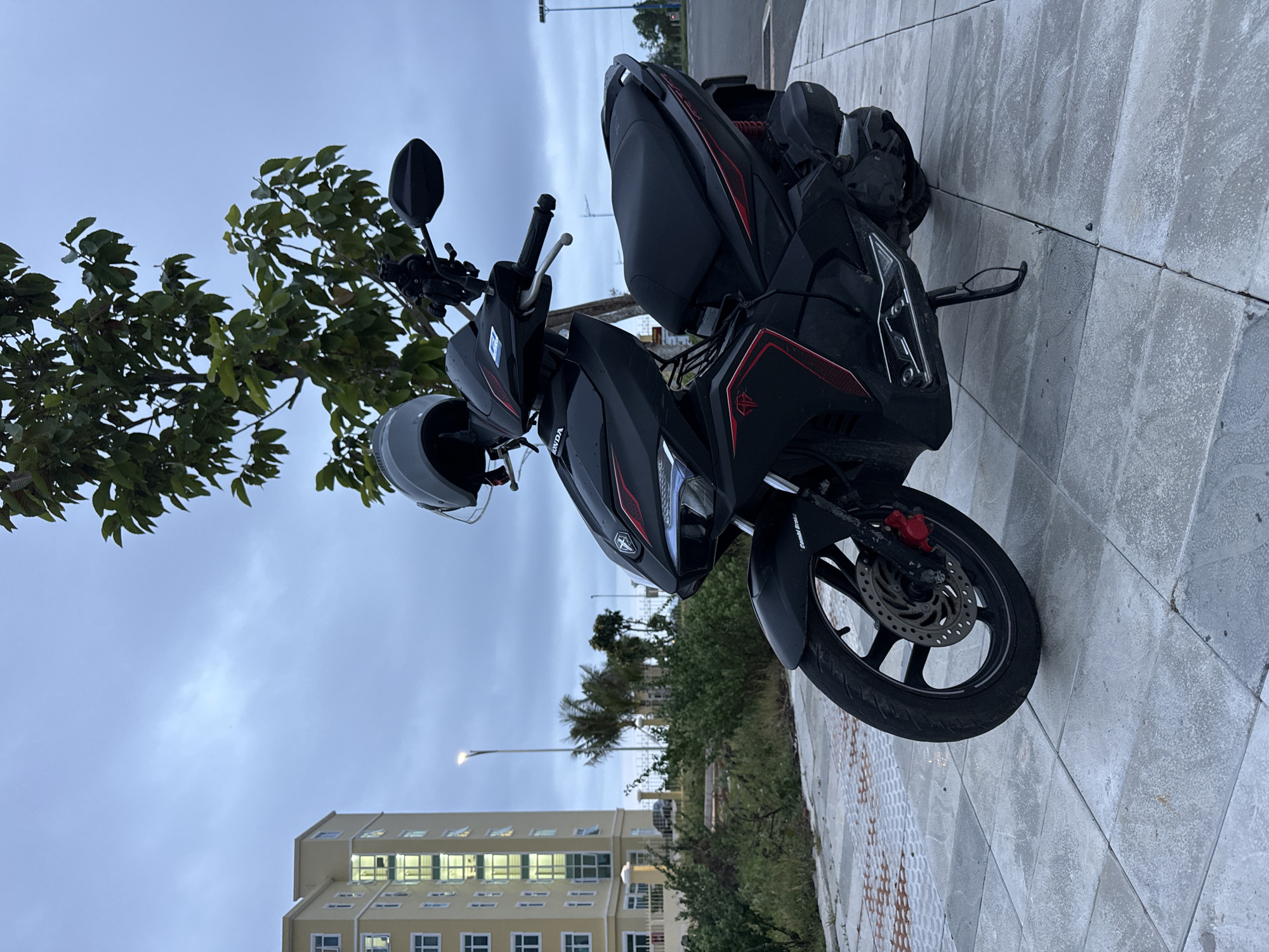An overcast sky looms above a black Honda motorcycle parked on a tiled sidewalk. The motorcycle, featuring red accents and the Honda logo, has a gray helmet resting on its handlebars. To the left of the motorcycle, a tall, light yellow building with numerous windows stands against the cloudy sky. A tree with green leaves is visible behind the motorcycle. In the background, palm trees and a white fence line the area, suggesting a location near the coast. The scene appears to be in Mui Ne, Vietnam, a coastal town known for its beaches and resorts. The overall impression is one of a quiet, possibly early morning or late afternoon setting, with the overcast weather muting the colors and creating a serene atmosphere.