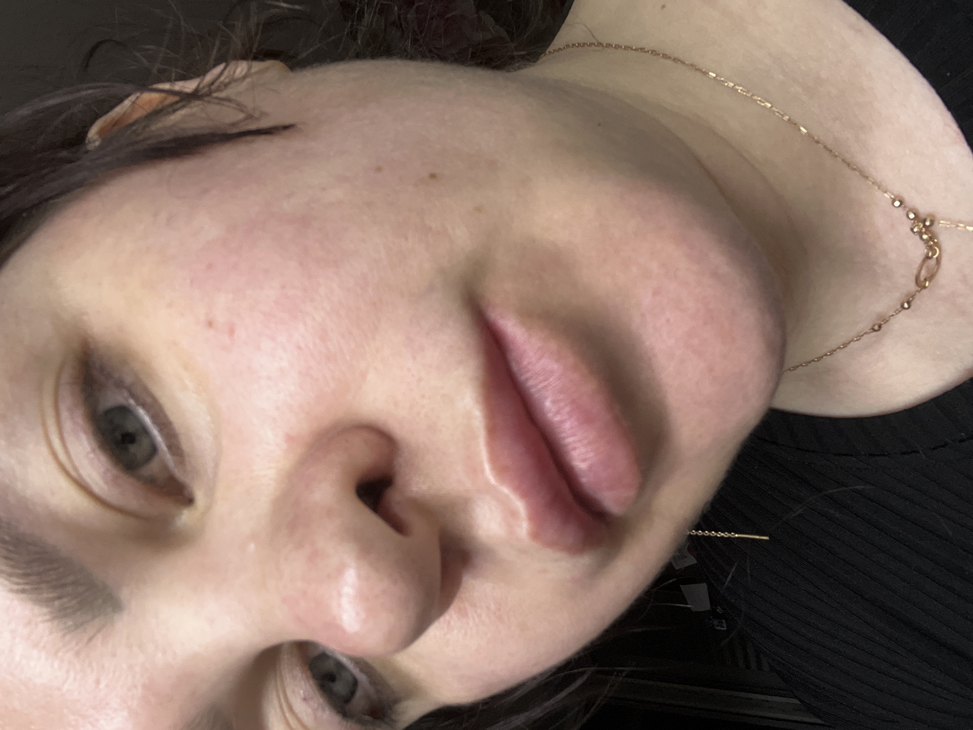 The image presents a detailed, close-up view of an adult female's face and upper chest, situated in the city of Noviy, Russia. Her fair skin shows localized redness on the cheeks and around the nasal area, along with visible pores and several small moles. The eyes are greenish-grey, and the full lips are a natural light pink, slightly parted. Dark brown, wavy hair is visible along the right side of her face. She is wearing a black, ribbed garment. A thin, gold-colored chain necklace with small beads and a visible clasp is present on her neck. A gold-colored threader earring, consisting of a chain and a small bar, hangs from her left earlobe. The background is uniformly dark and out of focus, indicating an indoor environment with minimal discernible features.