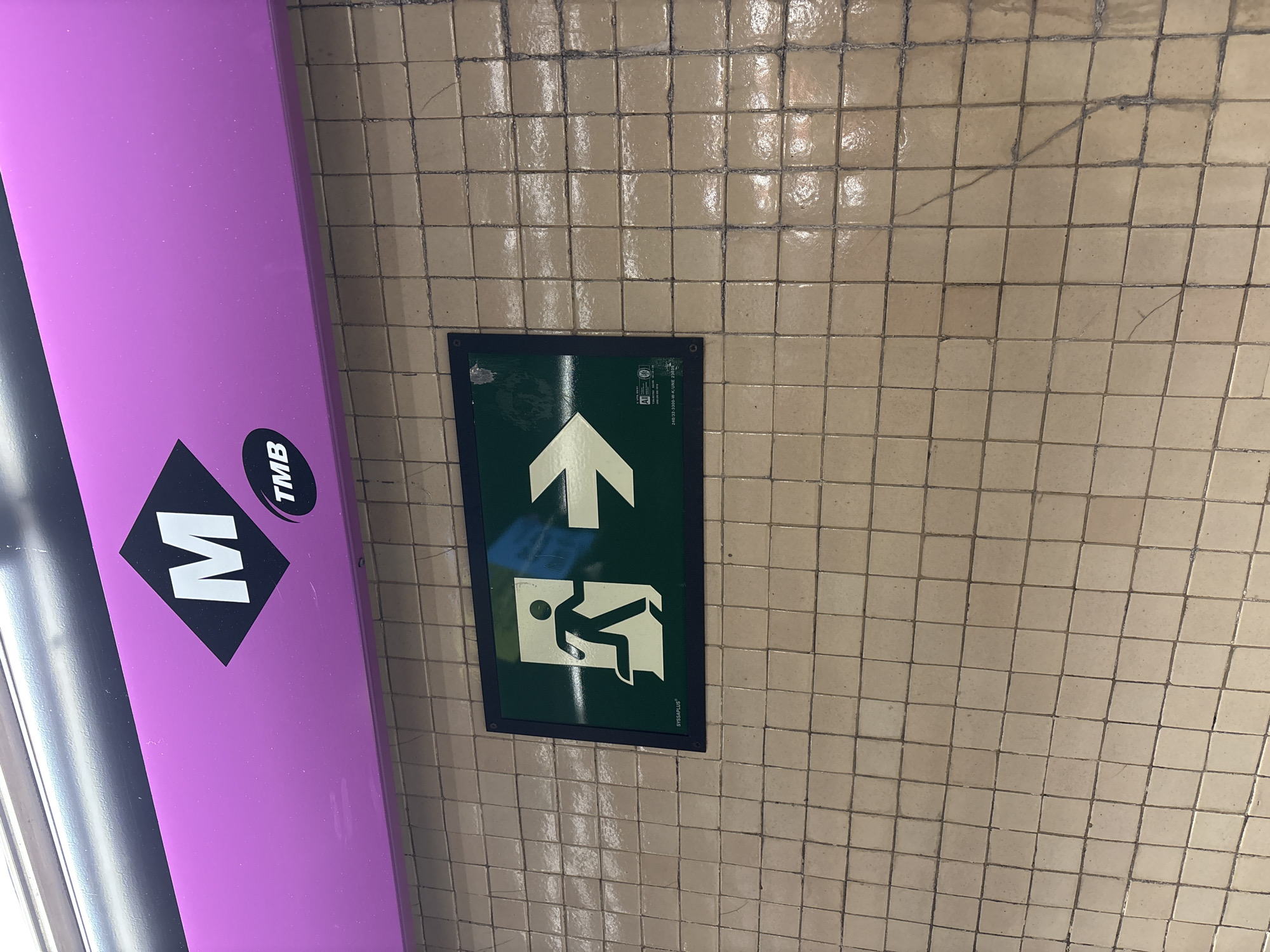 An overhead view shows a purple sign with a black diamond shape containing the letter "M" in white. To the right of the diamond, the letters "TMB" are in white within a black oval. Below this sign, a green rectangular sign with a white running figure and an arrow indicates an exit. The signs are mounted on a tiled wall, which appears to be in a subway or metro station. The tiles are light brown and square, with dark grout lines. The background is a blurred light.
