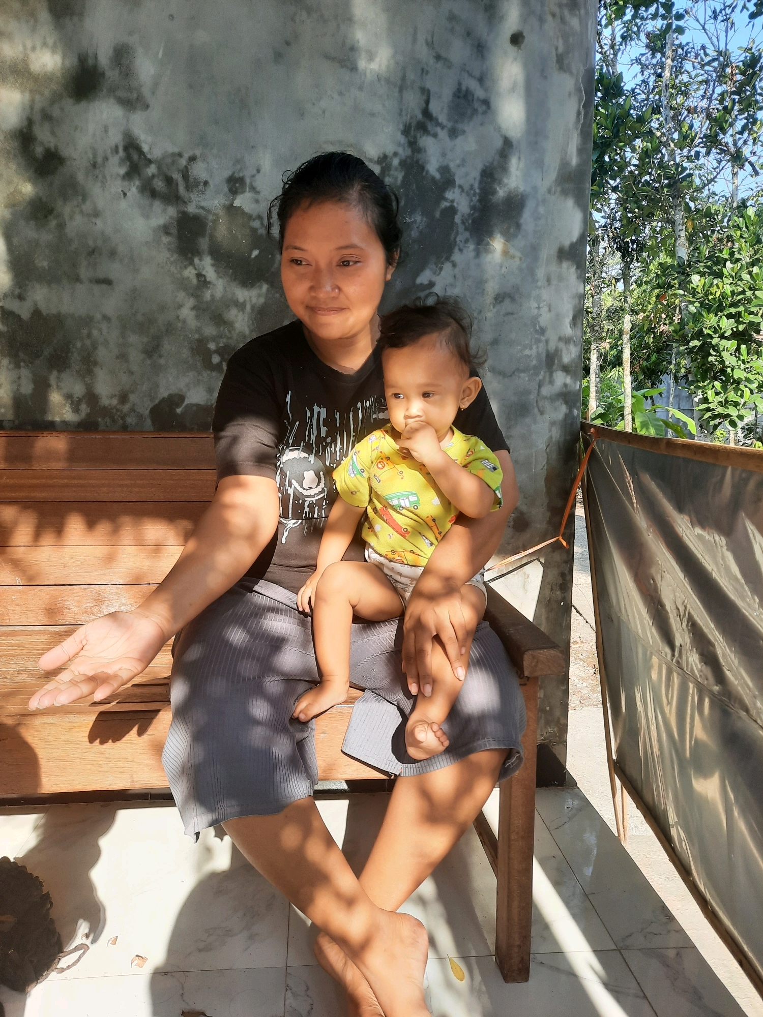 A person sits on a wooden bench, holding a baby. The person is wearing a black t-shirt and grey pants. The baby is wearing a yellow shirt and a diaper. The background is a concrete wall and trees. Sunlight streams in, casting shadows on the ground. The scene appears to be outdoors, possibly in a rural area, consistent with a location in Lugu, Indonesia.