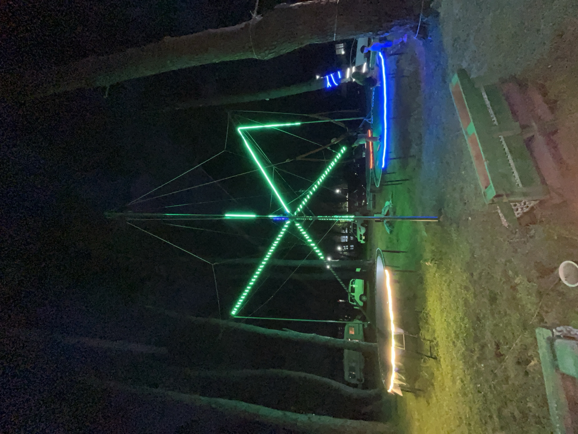 In the city of Ureki, Georgia, at night, an outdoor recreational area is illuminated by various lights. The central feature is a large, geometric structure made of metal poles and wires, adorned with bright green LED strips forming a star-like pattern. Two trampolines are set up on the grass. One, in the foreground, is circled with white string lights. The second trampoline, further back and to the right, features a bright blue LED strip around its edge. Several people are present, with two individuals visible on the blue-lit trampoline. In the background, several vehicles, including what appear to be RVs and cars, are parked. The area is surrounded by tall, dark trees. The ground is covered in grass, with some dirt and debris visible. A long, green wooden bench-like structure is positioned in the lower right foreground.
