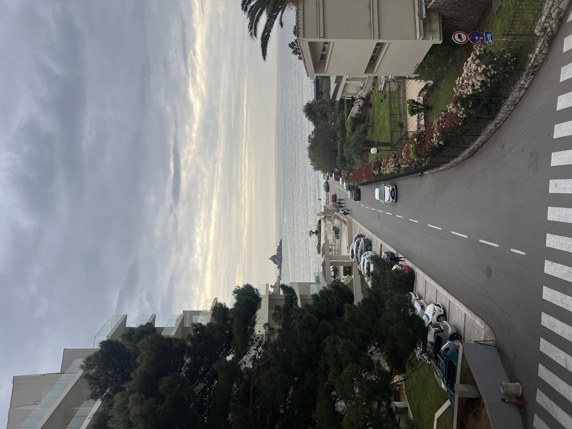 The scene depicts a street in Petrovac centar, Montenegro, with a view towards the Adriatic Sea. The sky is overcast with striated clouds, allowing diffused light. Buildings flank the street on both sides, displaying a mix of architectural styles. On the right, a multi-story building with balconies overlooks a green space bordered by flowering bushes. A road sign is visible at the street corner. A single white car travels down the street.
On the left, modern architecture is visible, featuring staggered balconies with glass railings partially obscured by trees. The street is lined with parked cars. Pedestrians can be seen in the distance near the waterfront. A small rocky island is visible in the sea. The road markings include a central dashed line and a pedestrian crosswalk in the foreground.