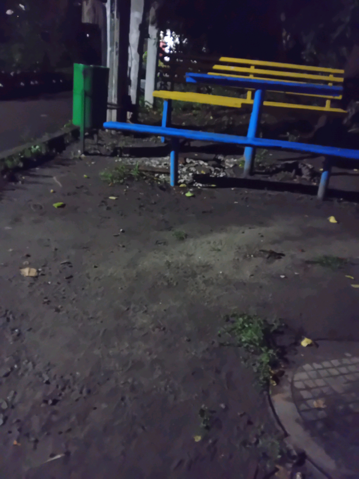 The image depicts an outdoor scene at night, possibly in a park or public space in Chuhuiv, Ukraine. A bright green trash can stands to the left. In the center, a public bench with yellow slats and a blue frame is visible. The ground is dark and appears to be paved, with scattered leaves and debris. A circular metal grate is partially visible in the lower right corner of the image. The lighting suggests artificial illumination from nearby sources, casting shadows. The overall environment appears somewhat unkempt.