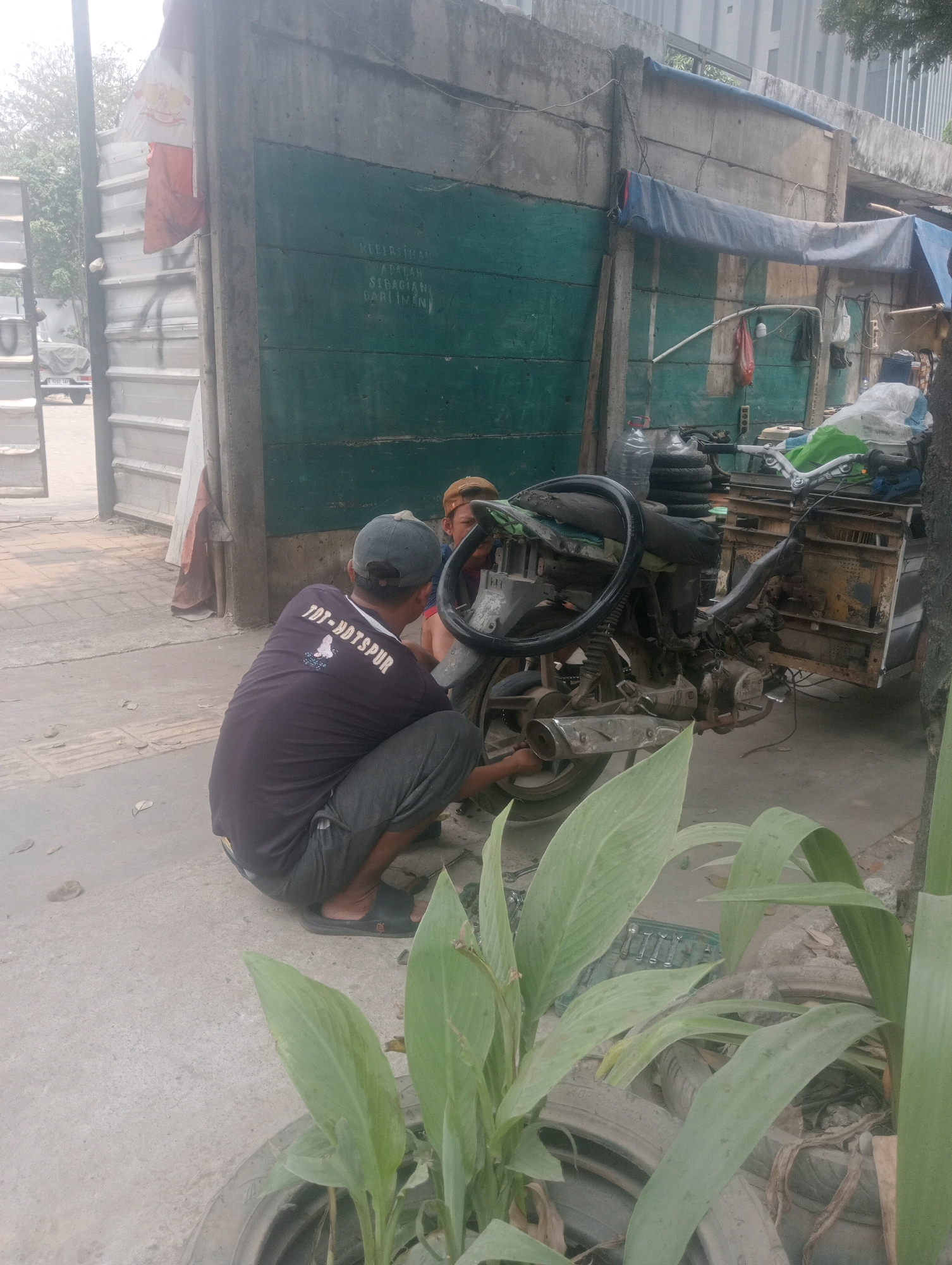 In South Jakarta, Indonesia, two individuals are engaged in repairing a three-wheeled motorcycle. One person, wearing a dark t-shirt with white lettering that reads "TOT-HDTSPUP" and grey pants, is kneeling down and appears to be working on the rear wheel of the motorcycle. The other person, wearing a brown cap and a reddish shirt, is also near the motorcycle, possibly assisting or observing. The motorcycle itself is weathered and consists of a front motorcycle section with a steering wheel and an engine, attached to a cargo bed at the rear. The surrounding environment is urban, with a concrete wall painted dark green in the background, adorned with white lettering. There are various materials and debris scattered around, including tires and tarpaulin coverings, suggesting a potentially informal or roadside repair setting. Sunlight illuminates the scene, casting shadows on the ground.
