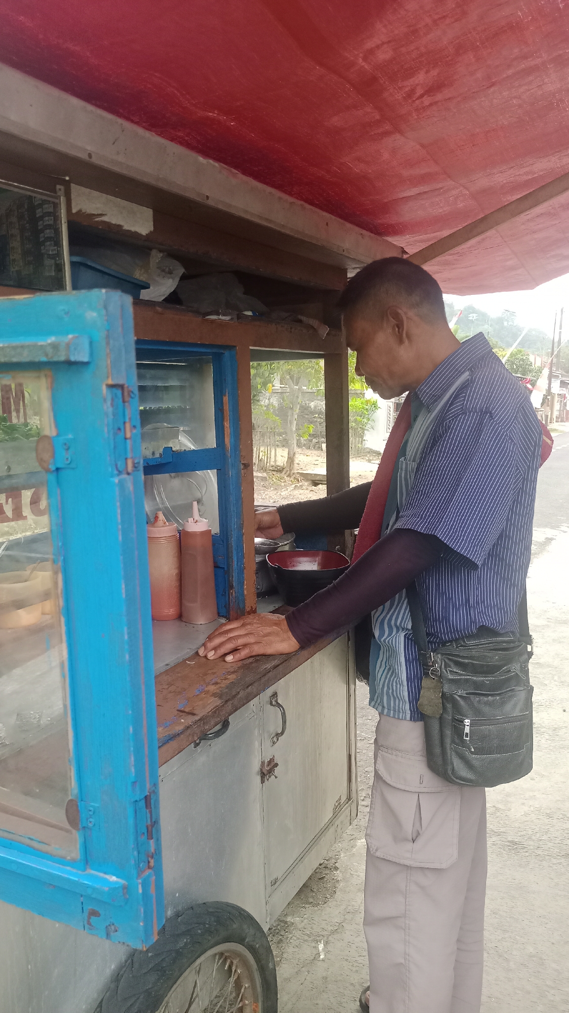 In Pakemitan, Indonesia, a man is standing behind a mobile food cart. He is wearing a blue and white striped short-sleeved shirt and light-colored cargo pants. A dark crossbody bag hangs from his right shoulder. He has dark skin and short, dark hair. His left hand rests on the wooden counter of the food cart, while his right hand appears to be reaching into the cart.
The food cart has a blue-painted wooden frame around a glass display case. Inside the case, there are two squeeze bottles, one with a red sauce and the other with a lighter brown sauce, and other food items. To the right of the display case, a red bowl is visible. The cart has a metal body and is supported by a wheel. A red awning is stretched over the top of the cart. The ground surface appears to be dirt or asphalt. In the background, there are trees and a building with a red and white flag.