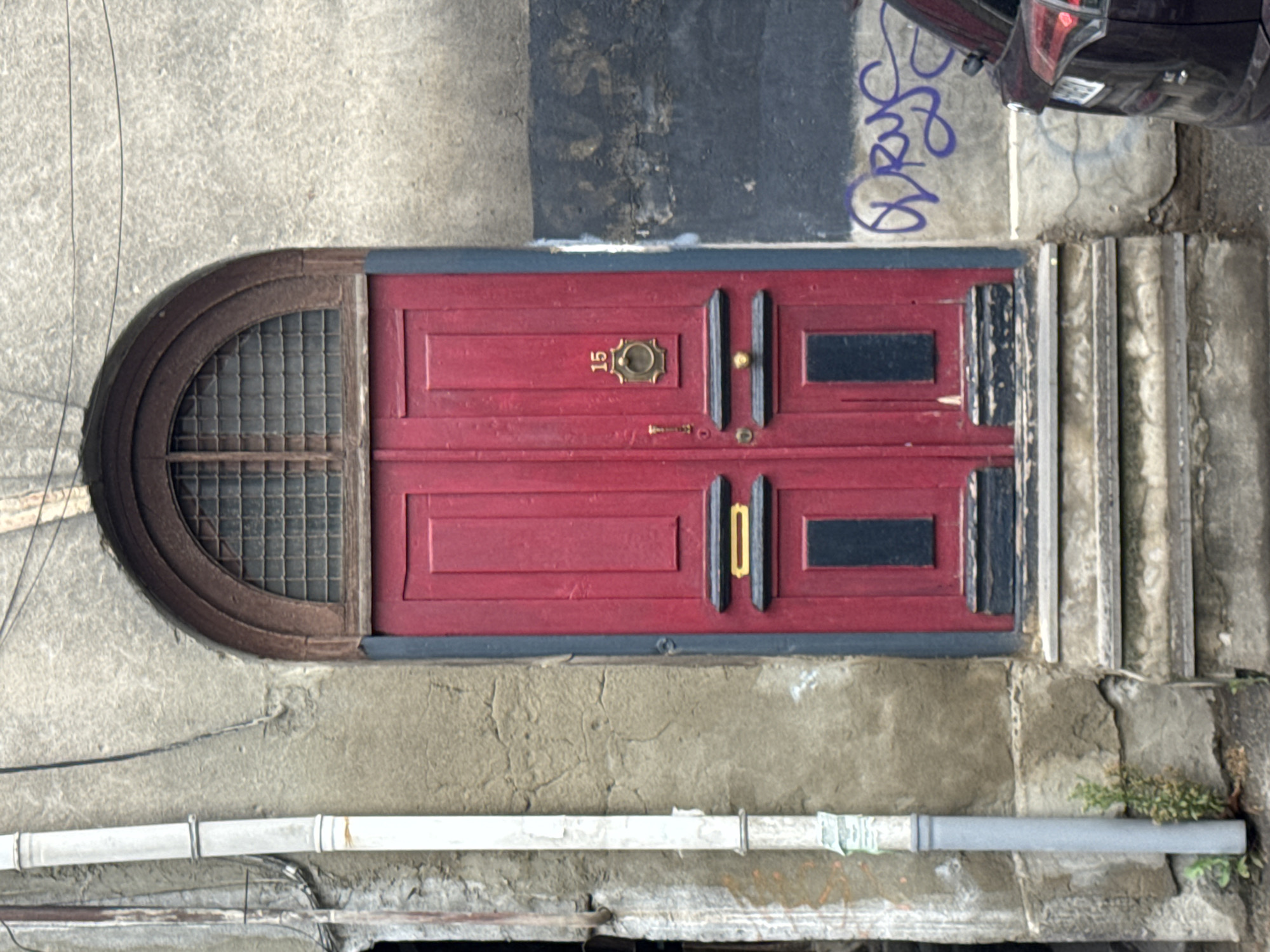 This is a medium shot, eye-level, outdoor photograph of an arched, red double door in Tbilisi, Georgia. The door is flanked by a textured, grey concrete wall on the right and a rough, grey stone wall on the left. A white downspout runs vertically along the left wall. The door has decorative panels, a brass door knocker with the number "15" above it, a brass handle, and a mail slot. Above the door is a semi-circular window with a grid of metal bars. The steps leading up to the door are made of weathered concrete. The wall to the right of the door has sections of black paint and graffiti. The rear of a dark-colored car is visible on the far right edge of the frame. The scene is overcast and the overall impression is one of age and a slightly dilapidated urban environment.