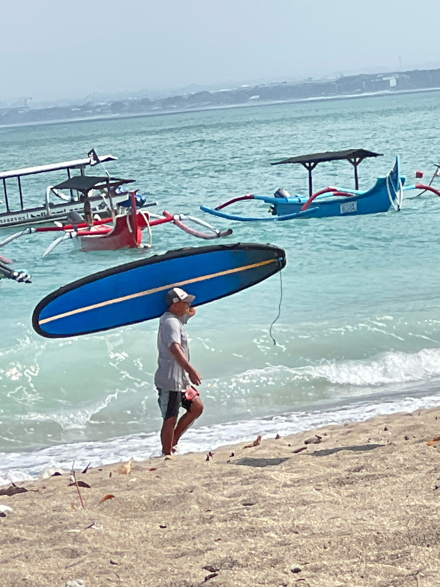 A man walks on the sandy beach in Kuta, Indonesia, carrying a blue surfboard on his right shoulder. The man is wearing a grey t-shirt, dark shorts with a red pattern, a white baseball cap, and white shoes. He is walking towards the right side of the image, with waves lapping at his feet. In the shallow turquoise water, several traditional Balinese jukung boats are anchored. One boat is painted bright blue with red outriggers and a canopy, and another is red with a white structure. The boats are bobbing gently in the water. The background shows a hazy coastline with buildings and a light grey sky. The sand is light brown and textured, with some debris scattered across it.