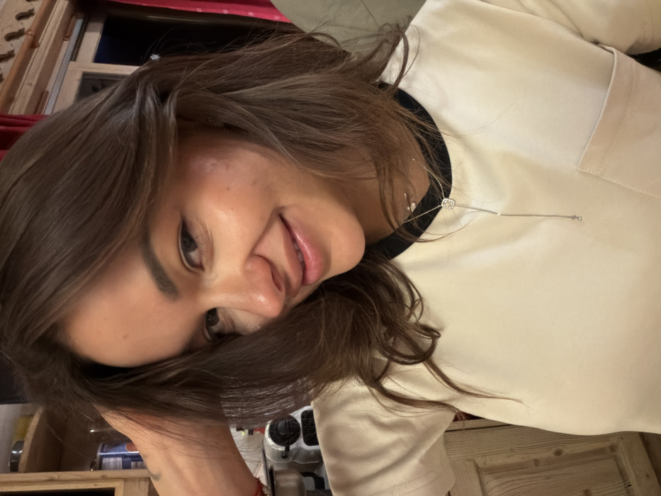 A woman tilts her head, smiling slightly towards the camera. She has long, brown hair that partially covers her face. Her skin appears smooth with minimal visible makeup. She is wearing a light-colored top with a dark trim at the neckline and a pendant necklace. The background features a mix of domestic elements like wood furniture, a red curtain, and what appears to be a stove or kitchen appliance. The lighting suggests an indoor setting.
