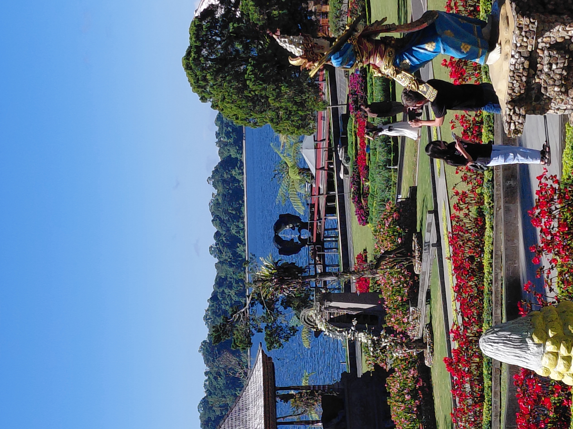 In Baturiti, Indonesia, a scenic view unfolds with a serene lake as the backdrop. In the foreground, a well-manicured garden bursts with vibrant red and pink flowers, interspersed with lush green grass. Several people are present, some capturing the scenery with their phones, while others are engaged in conversation. A statue of a traditional dancer stands prominently, adding a cultural touch to the scene. The architecture features a traditional Balinese gazebo with a thatched roof. The clear blue sky and the tranquil lake create a picturesque atmosphere.