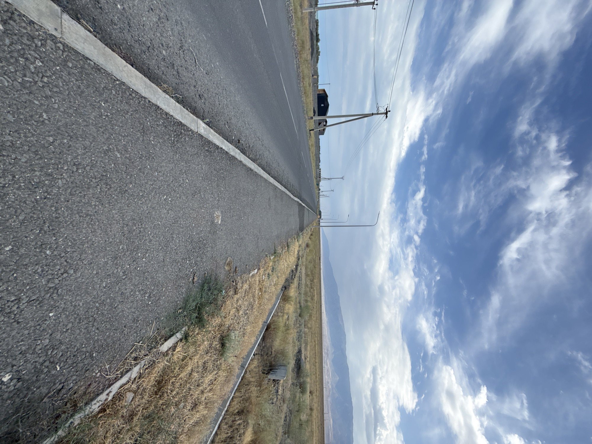 The image shows a view of a paved road and sidewalk running alongside a dry, grassy field. The road is asphalt and appears to be a two-lane road, with a white line marking the center. A concrete curb separates the road from the sidewalk, which is also paved. In the distance, there are mountains under a partly cloudy sky. Several utility poles with electrical wires are visible along the side of the road. In the background, a dark-colored building with a red roof is situated behind a fence. The overall scene suggests an outdoor environment in a semi-arid region, potentially on the outskirts of a town or city. The location in the city of Ақши, Kazakhstan, aligns with this observation, as it is situated in a region with a continental climate and mountainous terrain.
