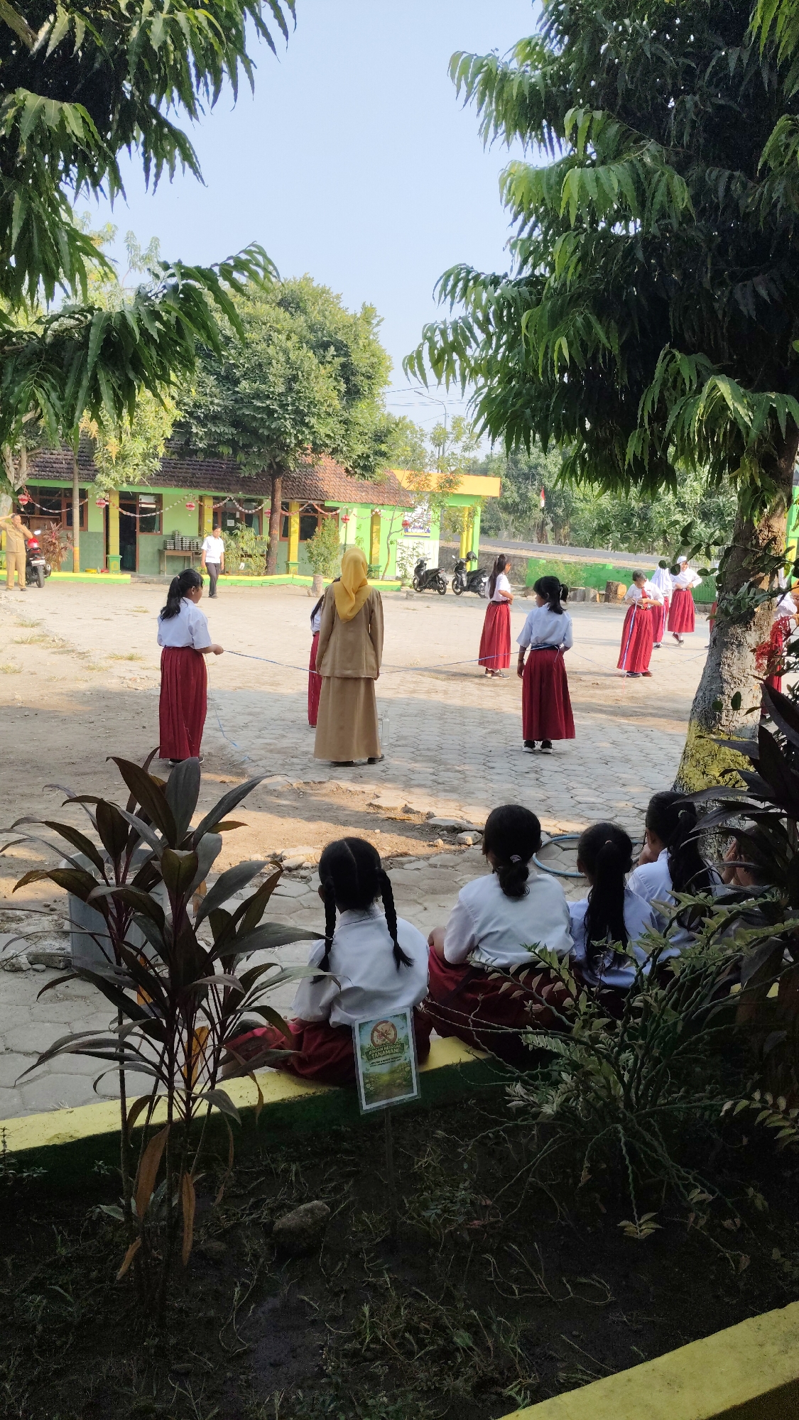 In the city of Banjarsari, Indonesia, a group of students in uniform, consisting of white shirts and long red skirts, are participating in an outdoor activity in what appears to be a schoolyard. The ground is a mixture of dirt and sand. In the background, a low-rise building with a green exterior and a brown roof is visible, suggesting it is part of the school infrastructure. Several trees with lush green foliage provide shade, creating a dappled sunlight effect on the ground. Some students are actively engaged in a game, possibly with a rope, while others are observing. In the foreground, a few students are seated, facing away from the camera, also appearing to be spectators. A small sign is placed in front of them. The overall scene depicts a sunny day with a light blue sky, characteristic of a tropical climate.