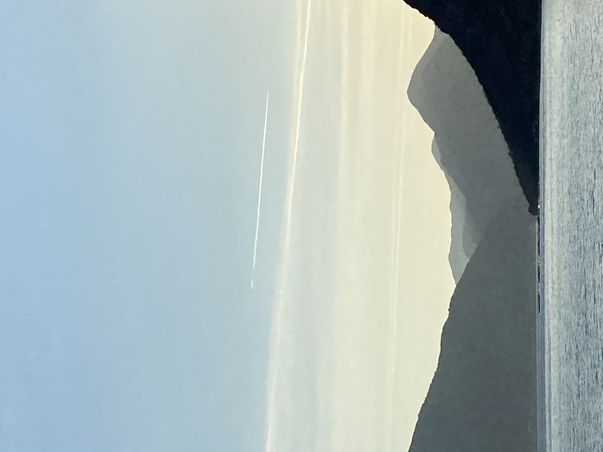 From Donji Orahovac, Montenegro, the image captures a view of layered mountains under a light sky. A vapor trail cuts vertically through the sky. A vertical wooden post is visible on the right side of the image.