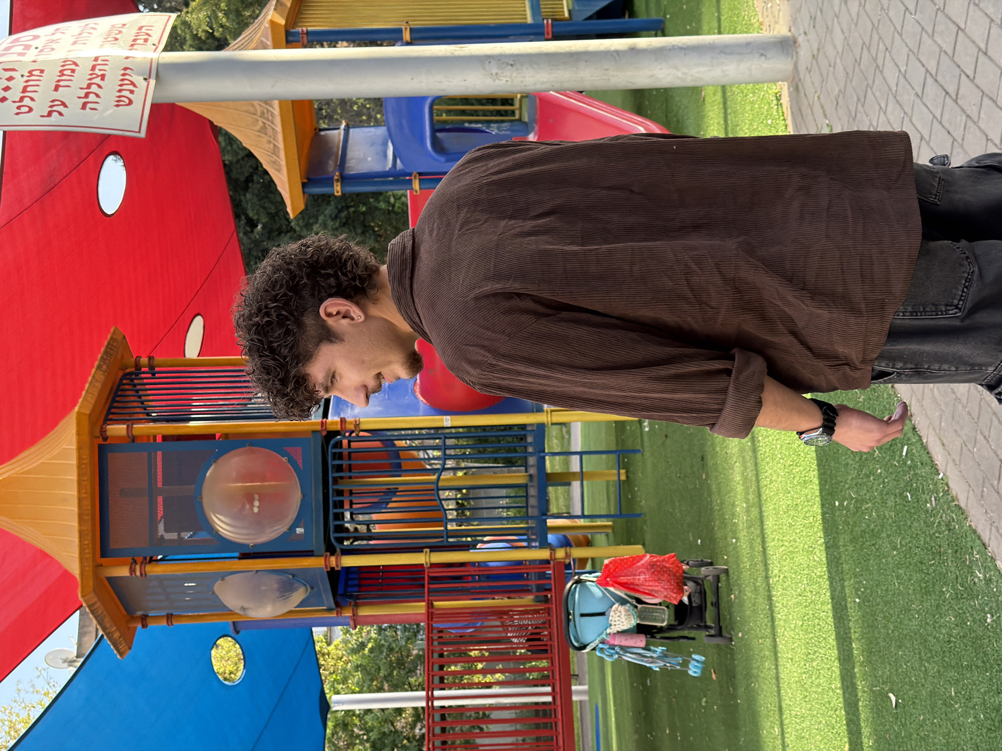 The image shows a man in a brown corduroy shirt and dark jeans standing next to a playground in Kiryat Bialik, Israel. The playground is brightly colored with red, yellow, blue, and green elements. A baby stroller is visible near the playground. The background includes green grass and trees.