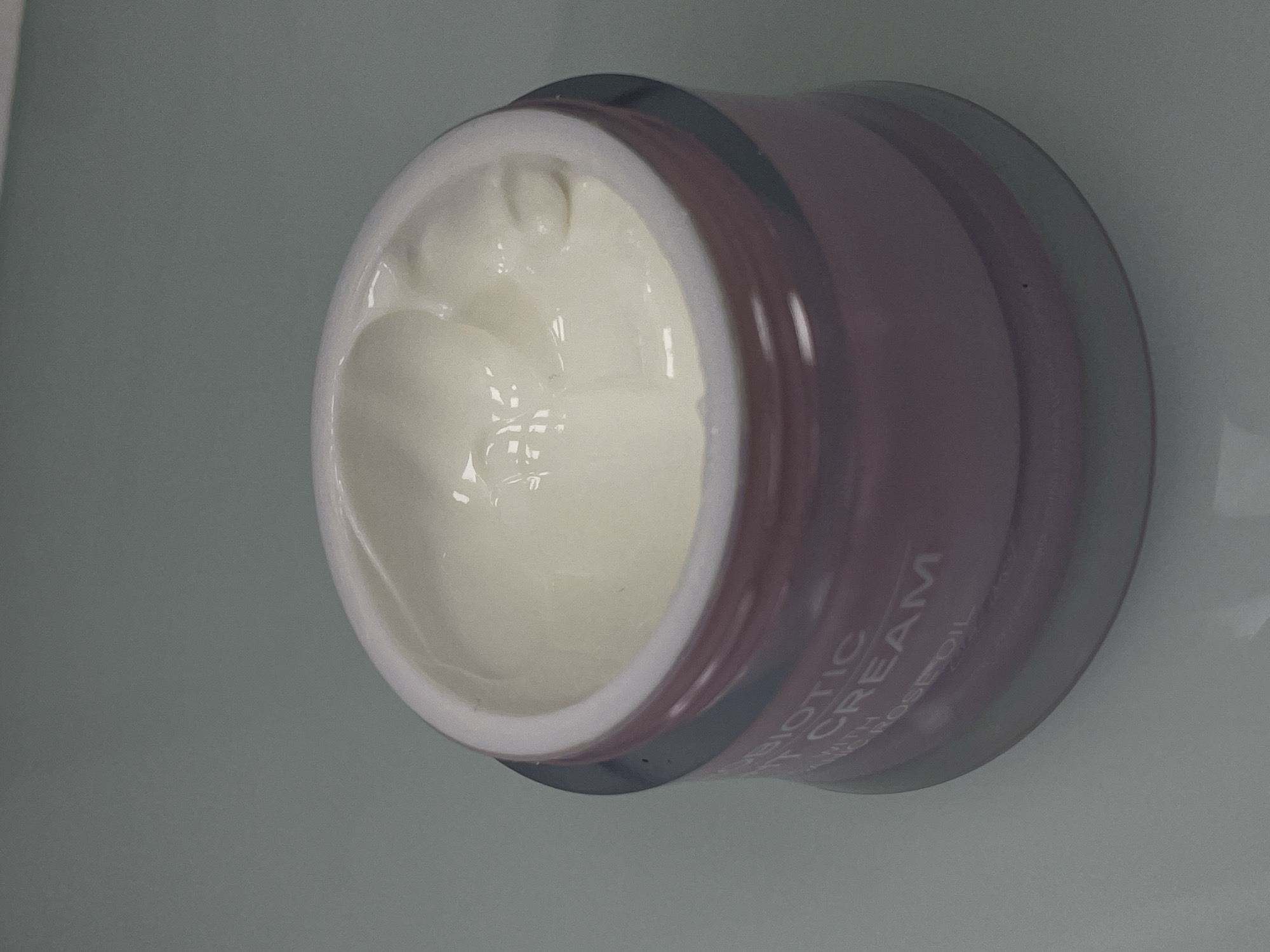 A close-up, slightly angled shot shows the top of an open jar of white cream on a gray surface. The jar has a pinkish-purple translucent band around its base with white text that reads "BIOTIC CREAM WITH ROSE OIL". The cream inside the jar is smooth and appears slightly glossy. The background is a uniform gray. There are no people or other objects visible in the image. The scene is likely indoors. The location of Gibraltar, Gibraltar is not depicted in this image.