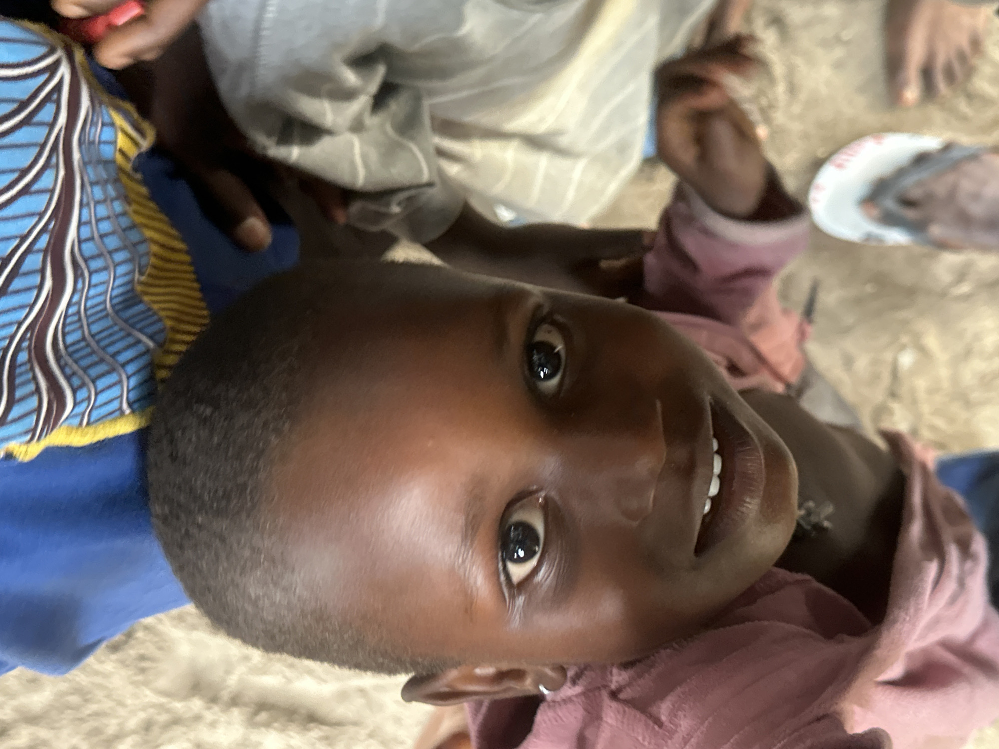 A close-up, eye-level shot features a young child in the foreground, with their face filling most of the frame. The child has dark skin, dark eyes, and a small smile revealing white teeth. They are wearing a pink shirt. Behind the child, a blue cloth with yellow and brown patterns is visible. To the right of the child, another person's arm is visible, wearing a grey and white striped garment. The background appears to be a sandy, outdoor environment, consistent with a location in Badagry, Nigeria. In the lower right of the frame, a flip-flop is visible, suggesting the presence of another person or the photographer.