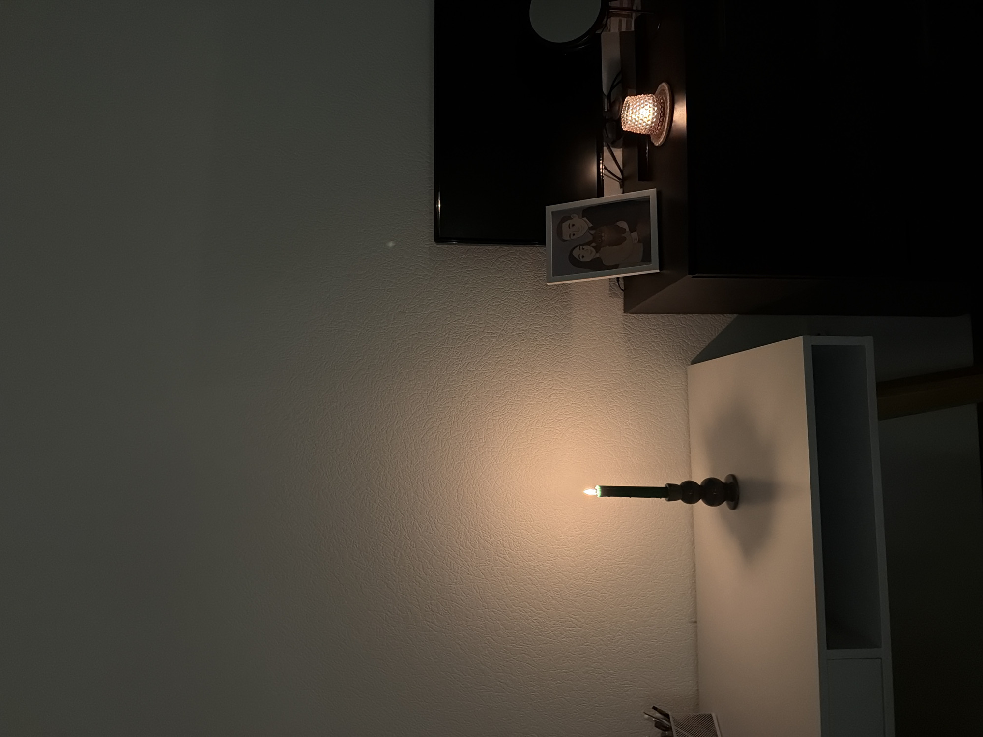 The image depicts a dimly lit interior scene, likely a bedroom or study, with a focus on a lit candle on a white desk. The candle, with a dark base and a green flame, casts a warm glow on the textured wall behind it. To the right of the candle, on a dark dresser, are several objects: a framed picture of a couple holding a pet, a small decorative lamp emitting a soft orange light, and a black television screen. A circular mirror is also visible to the right of the television. The overall atmosphere is somber and quiet. The location is unspecified and cannot be determined from the image.