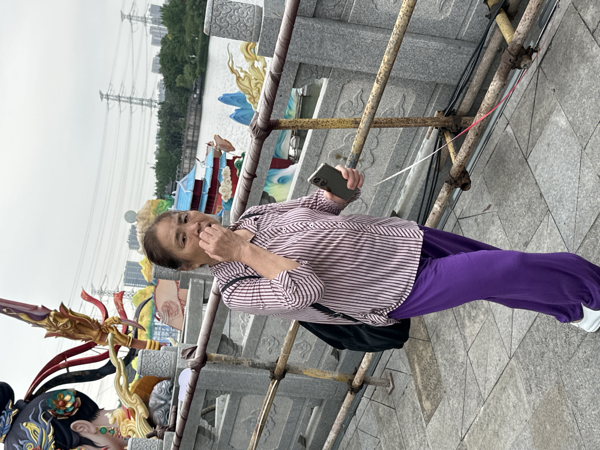An elderly woman stands on a paved surface in Liangjiang, China, holding a smartphone and looking at the camera. She wears a striped shirt and purple pants, with a black bag hanging off her shoulder. She's positioned in front of a stone railing and scaffolding, with decorative statues visible. A body of water, trees, and buildings are in the background. Electrical towers can be seen in the sky.