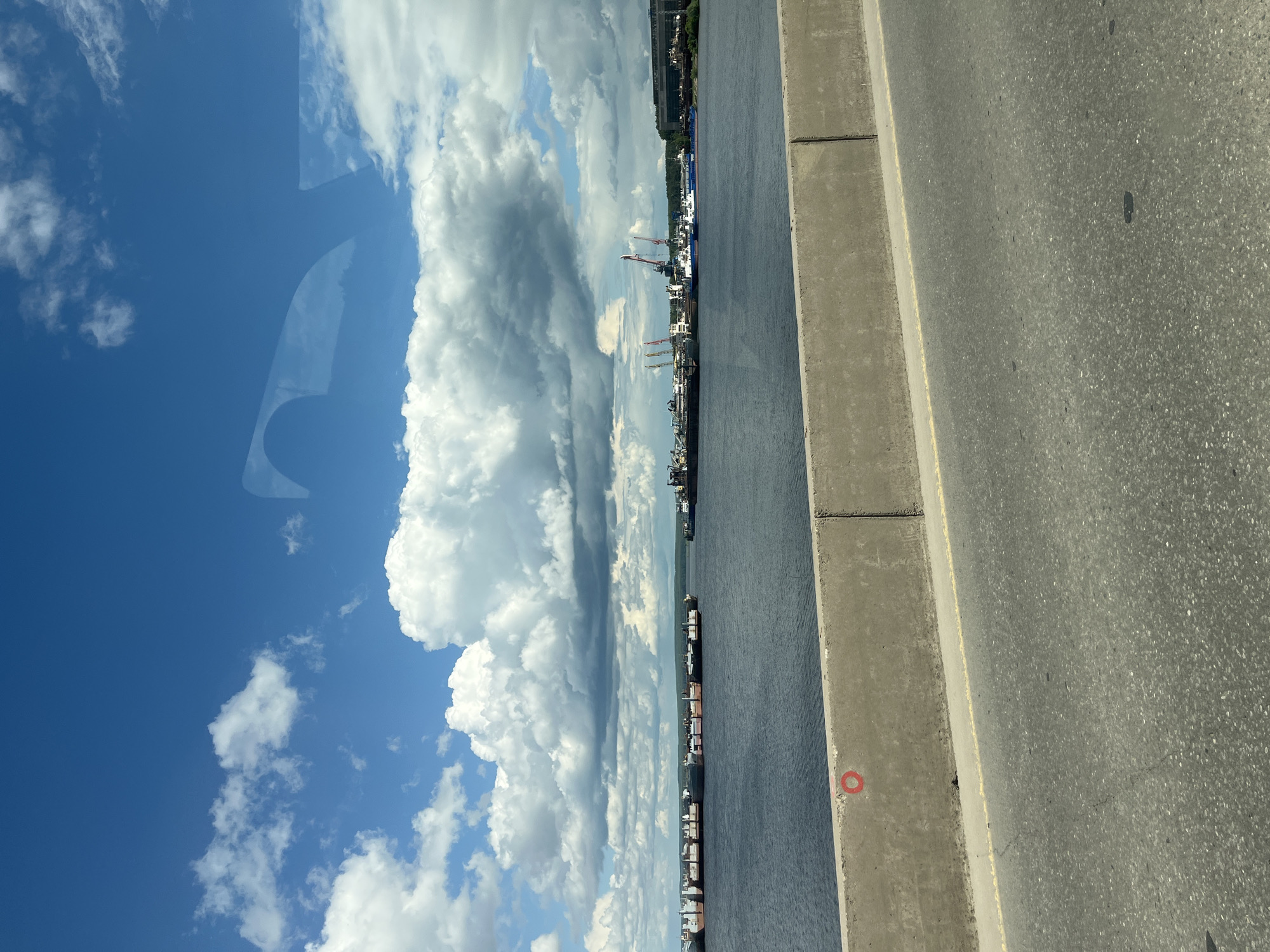 The image shows a view from a bridge in Chaikovsky, Russia, overlooking the Kama River. A line of cargo ships is docked along the shore. The sky is mostly blue with large, cumulus clouds. Part of the roadway and the bridge's concrete barrier are visible in the foreground. The image appears to have been taken through a car window, evidenced by a reflection on the left side of the frame.
