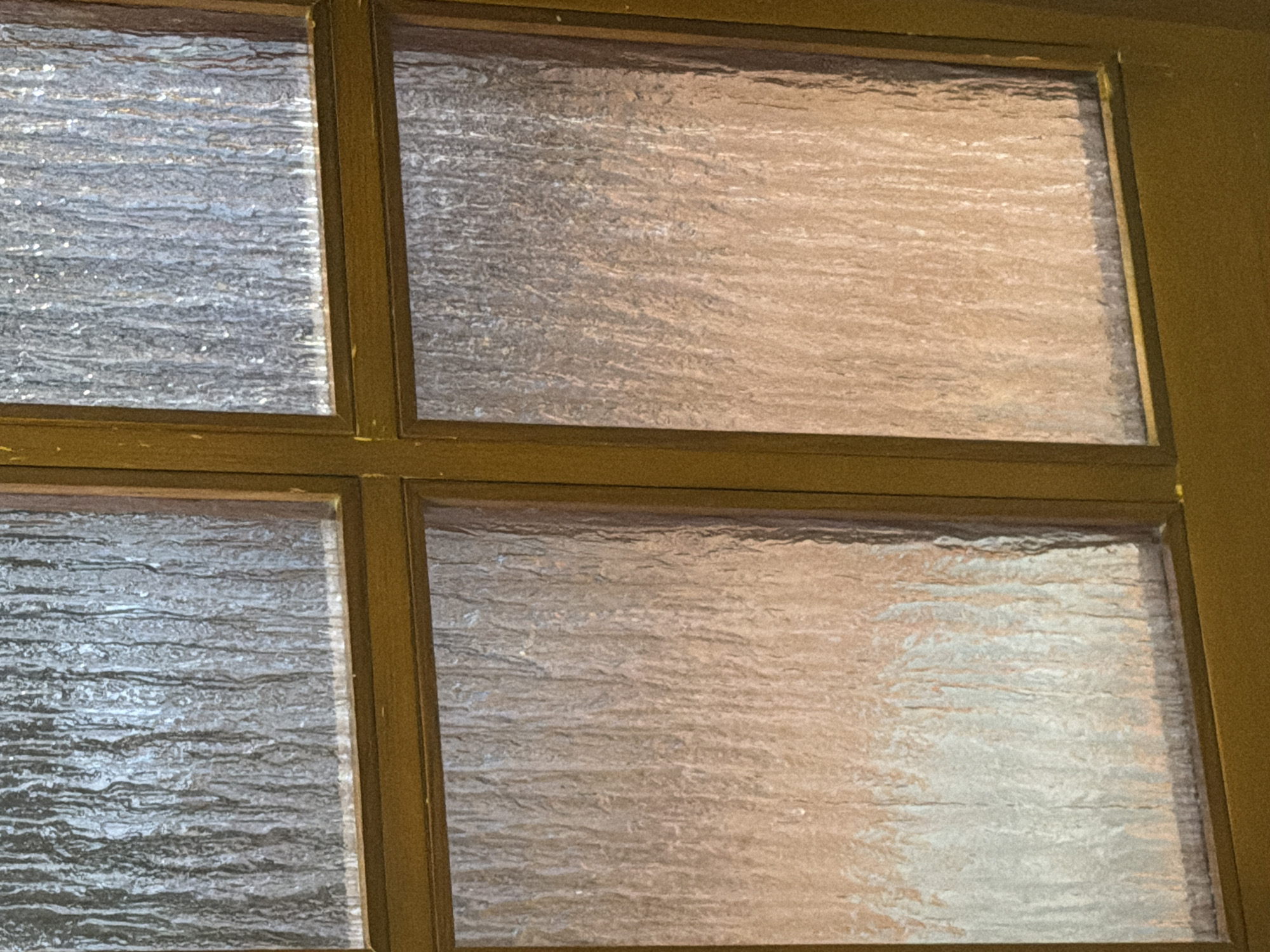 The image shows a close-up view of a multi-pane window with textured glass. The window frame appears to be made of wood and painted a dark, yellowish-brown color. The glass has a rippled or wavy texture, which distorts the view of what is behind it, creating abstract patterns of light and shadow. The lighting suggests it is either daytime with indirect sunlight or artificial light is illuminating the scene from behind the glass. The background visible through the glass has a warm, reddish-brown hue in some areas, and lighter, more muted tones in others, suggesting a wall or another room. There are no people or specific objects clearly identifiable through the textured glass. The image focuses solely on the window pane and its frame, with the textures and light playing a significant role in the visual composition. The image is not specific to Khmelnytskyi, Ukraine, and provides no contextual clues to suggest this location.