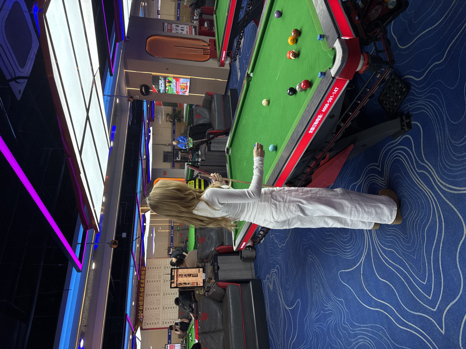 A woman in white plays pool in a brightly lit game room. The room features blue patterned carpeting and multiple pool tables. Other people are seated at gaming stations with monitors visible in the background. Bright, decorative lighting fixtures line the ceiling on one side of the room.