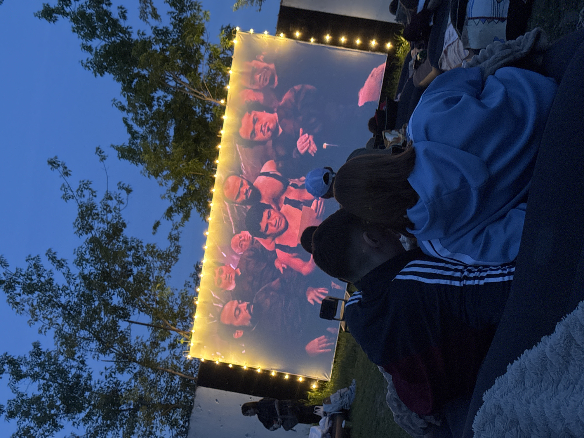 The image shows an outdoor movie screening taking place at night. A large white screen, illuminated by string lights, displays a film featuring a group of male actors. Several individuals are seated on the ground, watching the movie. Two people, one wearing a blue hoodie and another in an Adidas tracksuit, are prominently visible in the foreground. Trees are present on the left side of the frame, and the sky is a dark blue. The specific location within Saratov, Russia cannot be determined from the image.