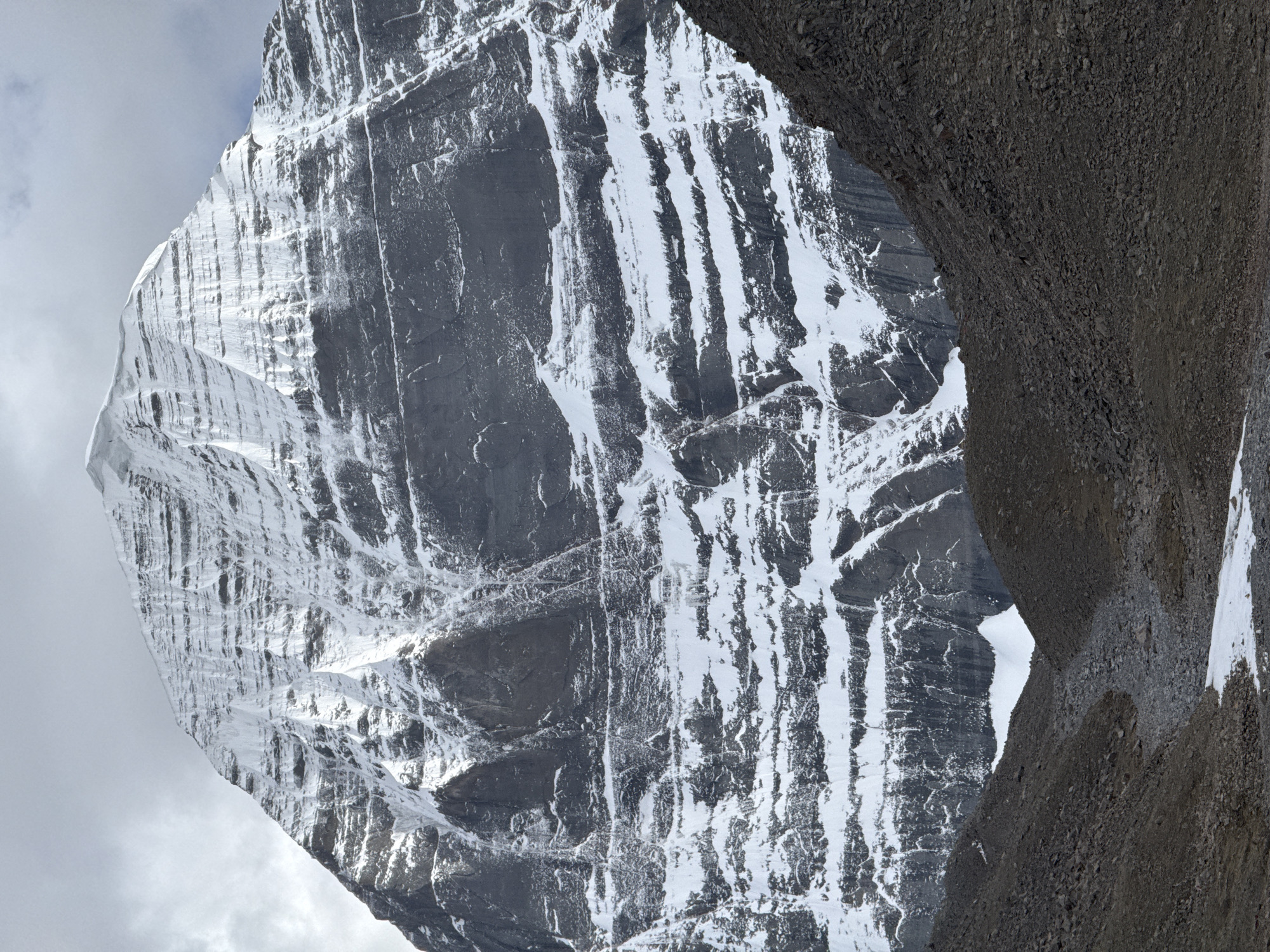 The image shows a large, gray mountain with snow-covered vertical streaks. The mountain is partially framed by a darker, rocky slope on the right. The sky behind the mountain is cloudy, with a gray hue. No people or signs of urbanization are visible in the image. The location, Parga town, China, is not supported by visual elements within the image.