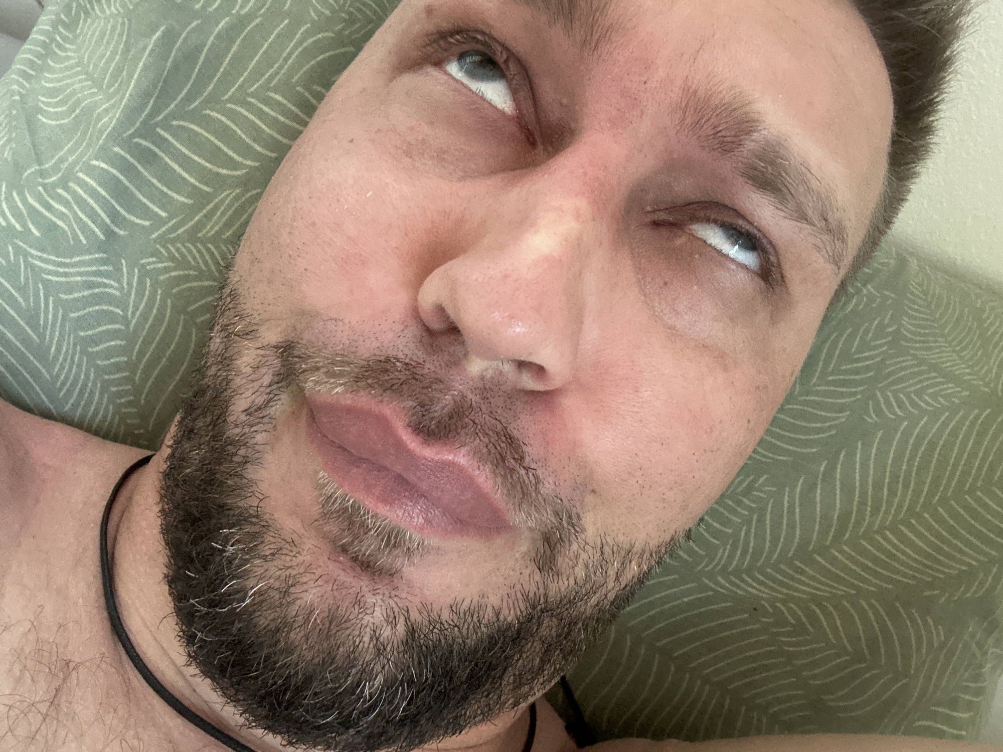 An extreme close-up, distorted, and low-angle shot focuses on the face of a man lying down, resting on a green textured surface that appears to be a pillow or bedding with a leaf-like pattern. The man's face, with light skin and a beard, occupies the majority of the frame. His eyes are open and looking upwards, with one eye more visible than the other. The lighting in the scene is soft and appears to be from above, casting subtle shadows on his face. The overall impression is intimate and candid, as if captured in a moment of relaxation or repose.