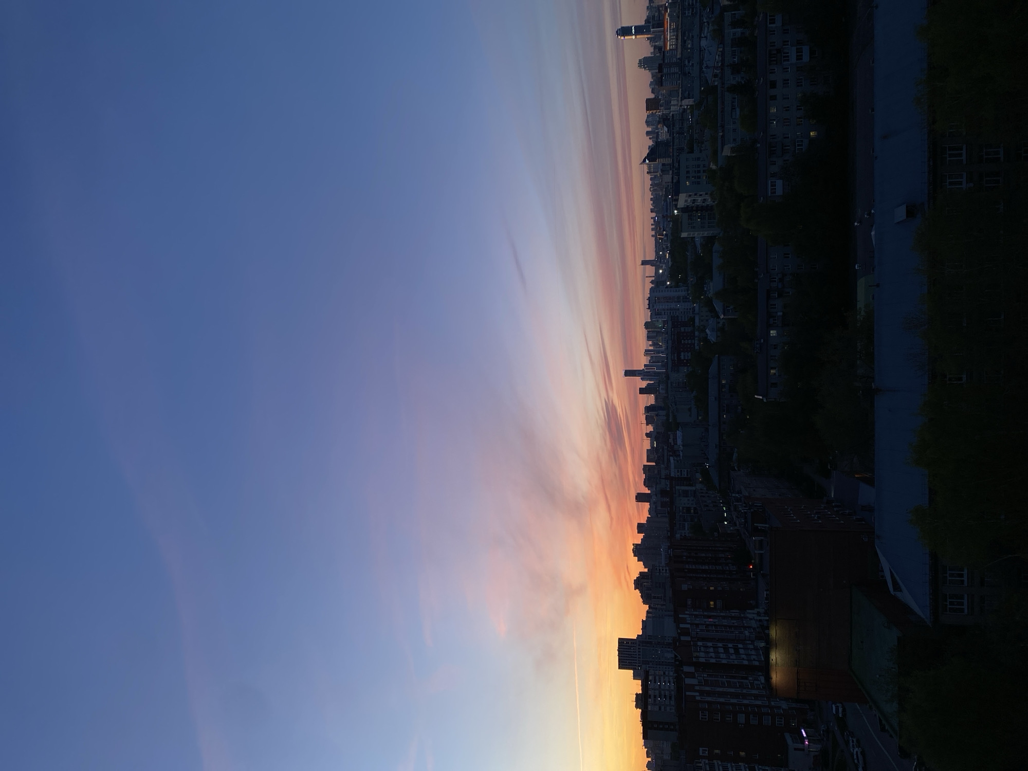 The image depicts a cityscape at dusk. The sky transitions from a deep blue at the top to shades of orange and pink towards the horizon. Wispy clouds are present. Below the sky, a dense urban landscape stretches across the frame. Buildings of varying heights are visible, some illuminated with lights. Trees are scattered among the buildings. The location is Yekaterinburg, Russia.