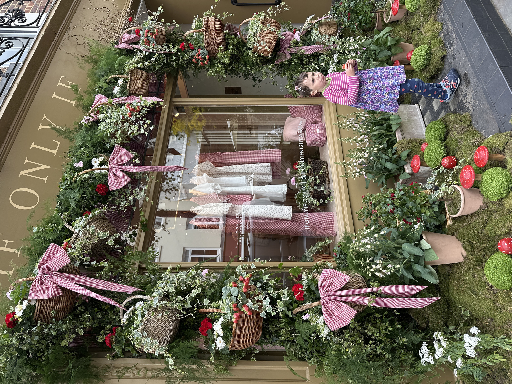 A young girl stands in front of a storefront in London, United Kingdom. The store window is decorated with an elaborate display of greenery, woven baskets, red gingham bows, red berries, white and red flowers, and artificial mushrooms. Inside the window, articles of clothing can be seen, including white and red-patterned garments. The street is paved with dark tiles, and the building facade is painted in neutral tones with white lettering.