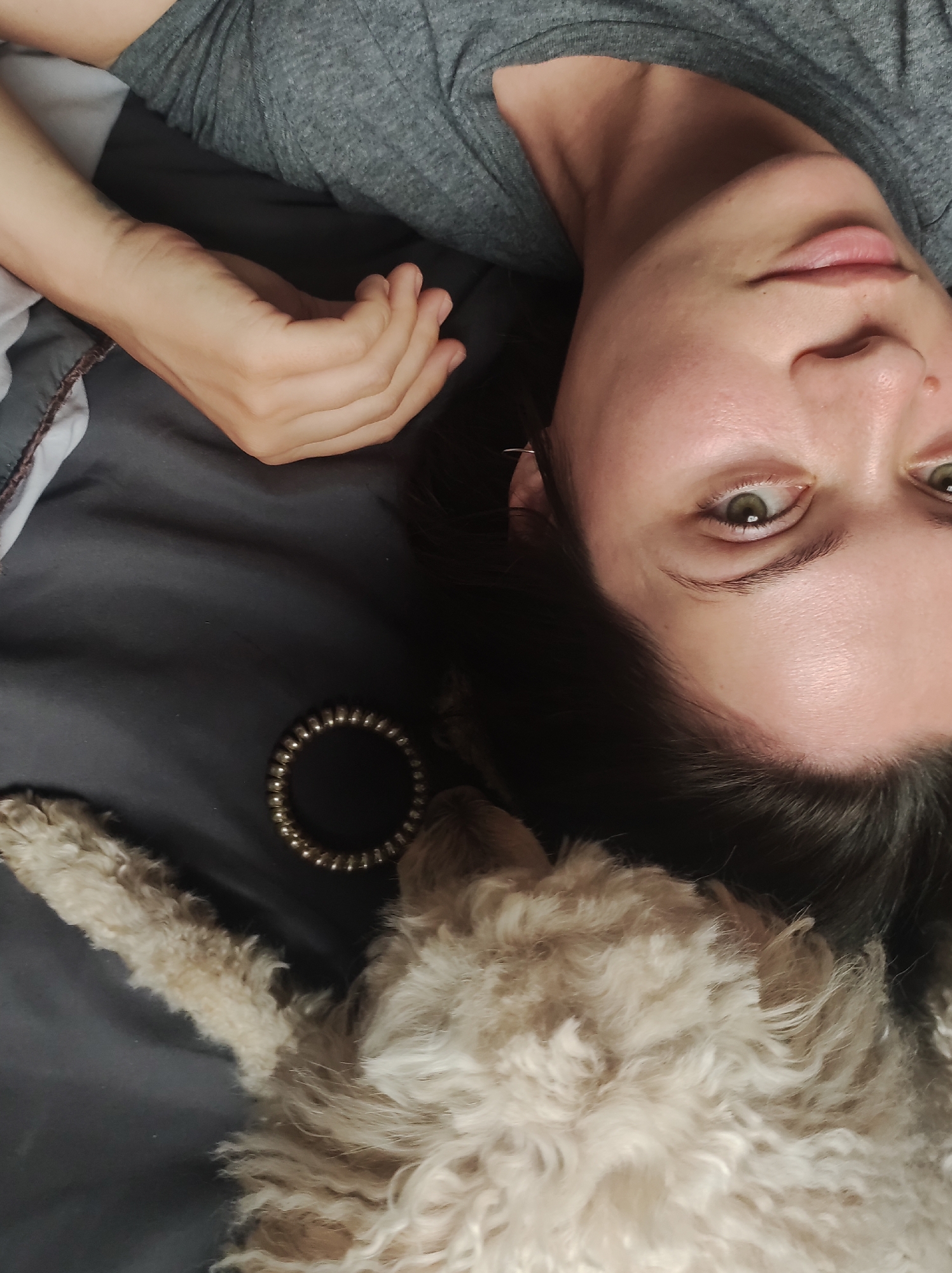 A woman lies on a gray surface, likely a bed. Her head is near the bottom center of the image, and she looks directly at the camera. She has dark hair and light eyes. A small, light-colored dog rests near her head. A beaded bracelet lies on the surface near the dog. The woman's hand is visible in the upper left portion of the image. She wears a gray shirt. The location is indeterminable. The city of Chaikovsky, Russia is also indeterminable from this image.