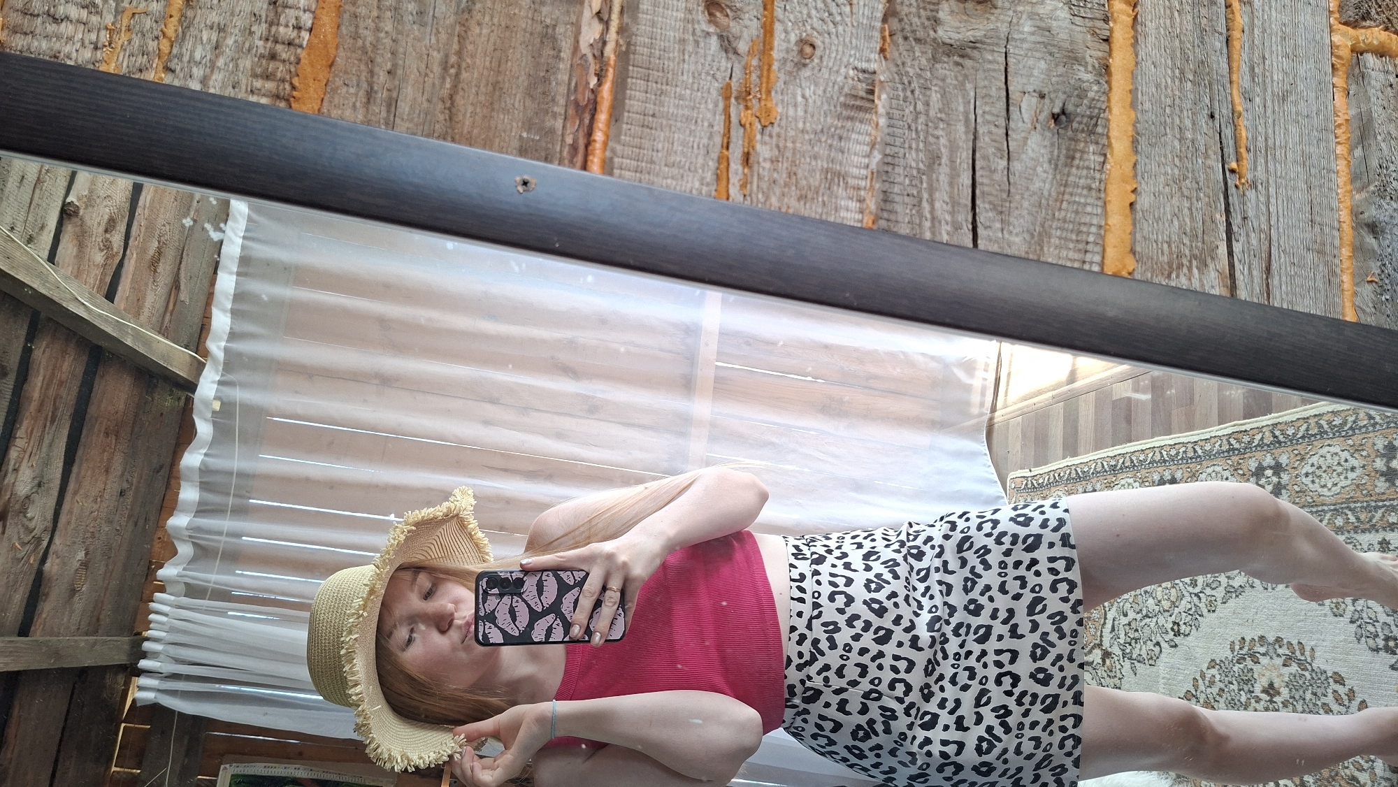 A reflection captured in a mirror shows a person wearing a sun hat, a red top, and a black and white animal print skirt. The person is holding a smartphone with a patterned case, presumably taking a selfie. The background reflected includes a window with a white curtain, wooden walls, and a carpeted floor. The mirror's frame is visible along the top edge of the image. The mirror is located in the city of Большой Букор, Russia.