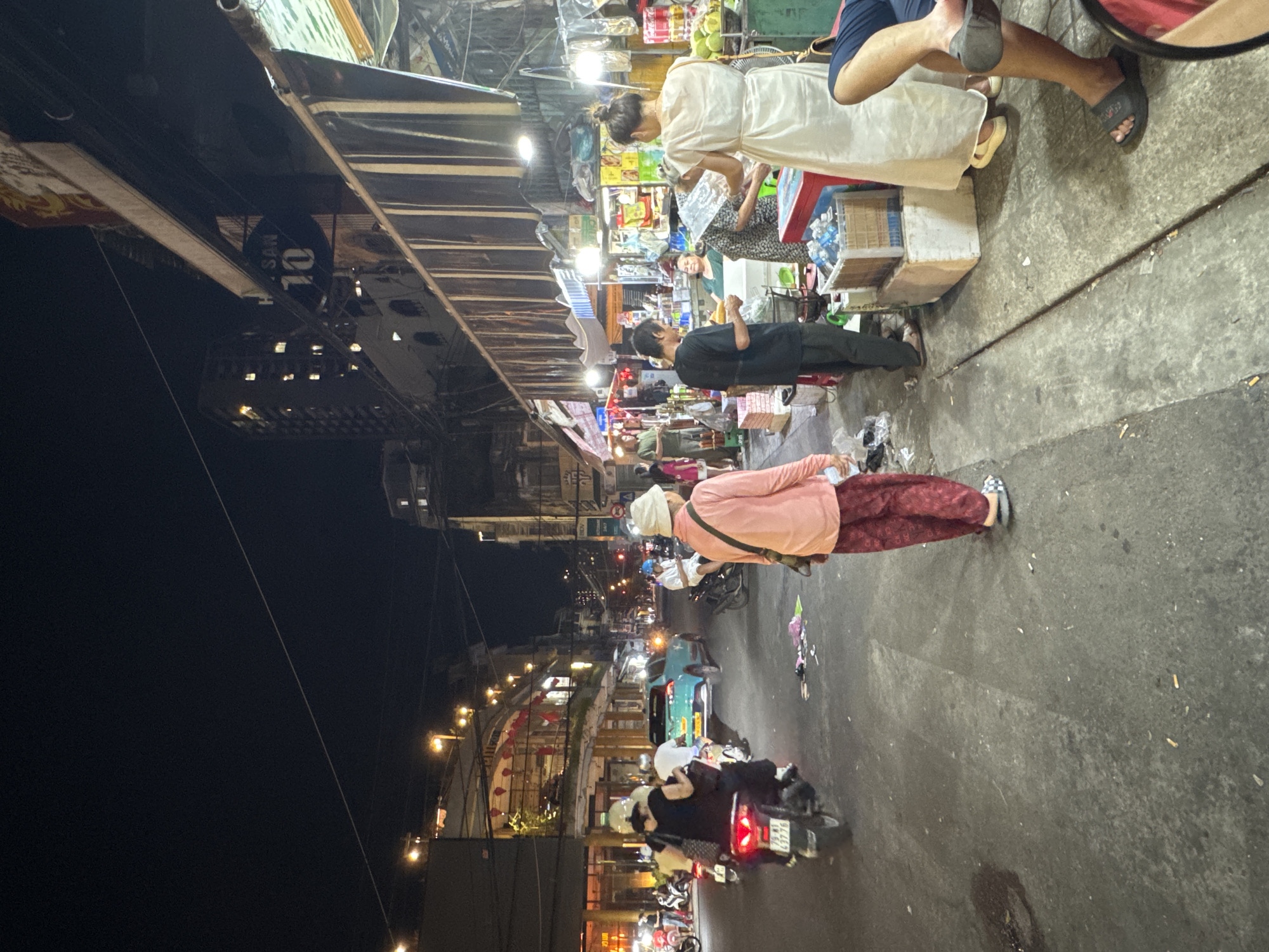 The image depicts a street scene at night in Nha Trang, Vietnam, likely a market or commercial area. A variety of people are present, including pedestrians and individuals on motorcycles.
In the foreground, on the right side of the image, a person wearing a blue garment is seated, their legs visible. To their left, a woman in a white dress stands near a market stall, appearing to interact with a man in a black shirt who is holding what seems to be a loaf of bread. Another woman in a patterned dress is partially visible behind them, and a person wearing a pale pink long-sleeved shirt and patterned pants stands in the center of the frame, facing away from the camera.
The street is paved and shows signs of wear. To the left of the frame, several motorcycles are parked or in motion. A turquoise car is visible further down the street. The background features buildings with illuminated windows, market stalls with various goods displayed, and overhead wires. Streetlights and shop lights illuminate the scene. The overall atmosphere suggests a bustling urban environment at night.