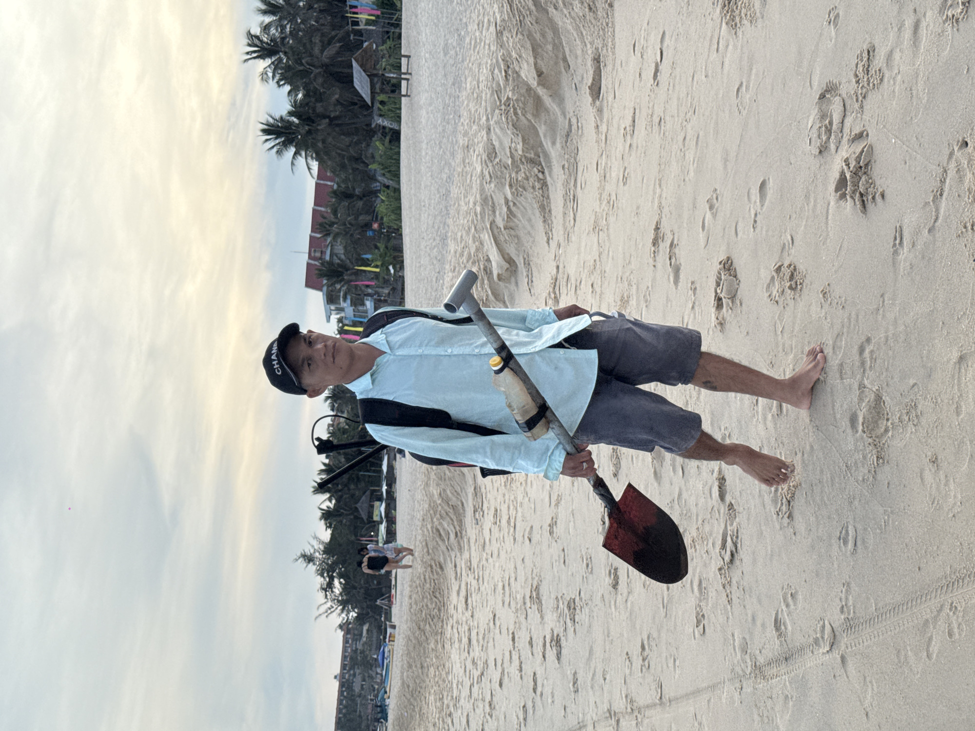 A man wearing a light blue long-sleeved shirt, dark shorts, and a black baseball cap stands on a sandy beach. He is carrying a metal detector on his back and a shovel in his right hand. The shovel has a red and black metal head and a gray handle. A plastic bottle is attached to the metal detector's pole. In the background, palm trees and buildings are visible. The sky is overcast. The location is Phu Thuy Ward, Vietnam.