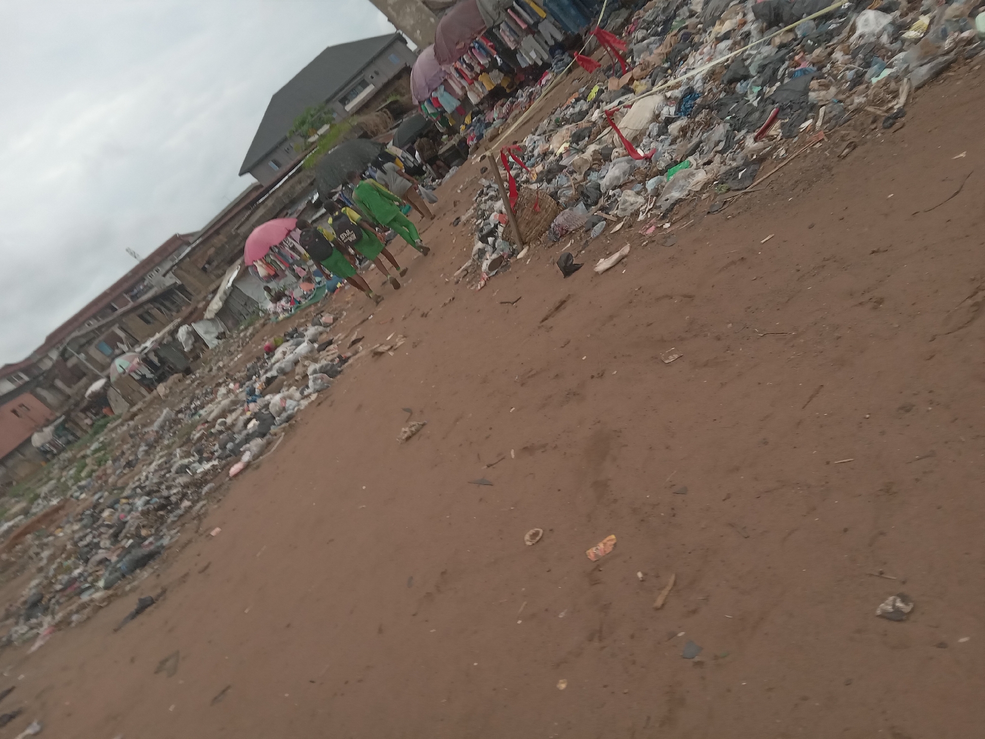 The image shows a street scene in Onitsha, Nigeria, characterized by significant environmental degradation and poverty. The ground is primarily brown soil, scattered with refuse and debris. Piles of mixed waste, including plastic, cloth, and unidentified materials, are visible along the roadside.
Several individuals walk along the left side of the image, some carrying umbrellas for protection from the weather. They are dressed in casual clothing, including school uniforms. The buildings lining the street are a mix of residential and commercial structures, appearing worn and dilapidated. Some have corrugated metal roofs and unfinished facades. Clothes hanging and for sale suggest a marketplace atmosphere. The sky is overcast, contributing to the muted color palette of the scene.