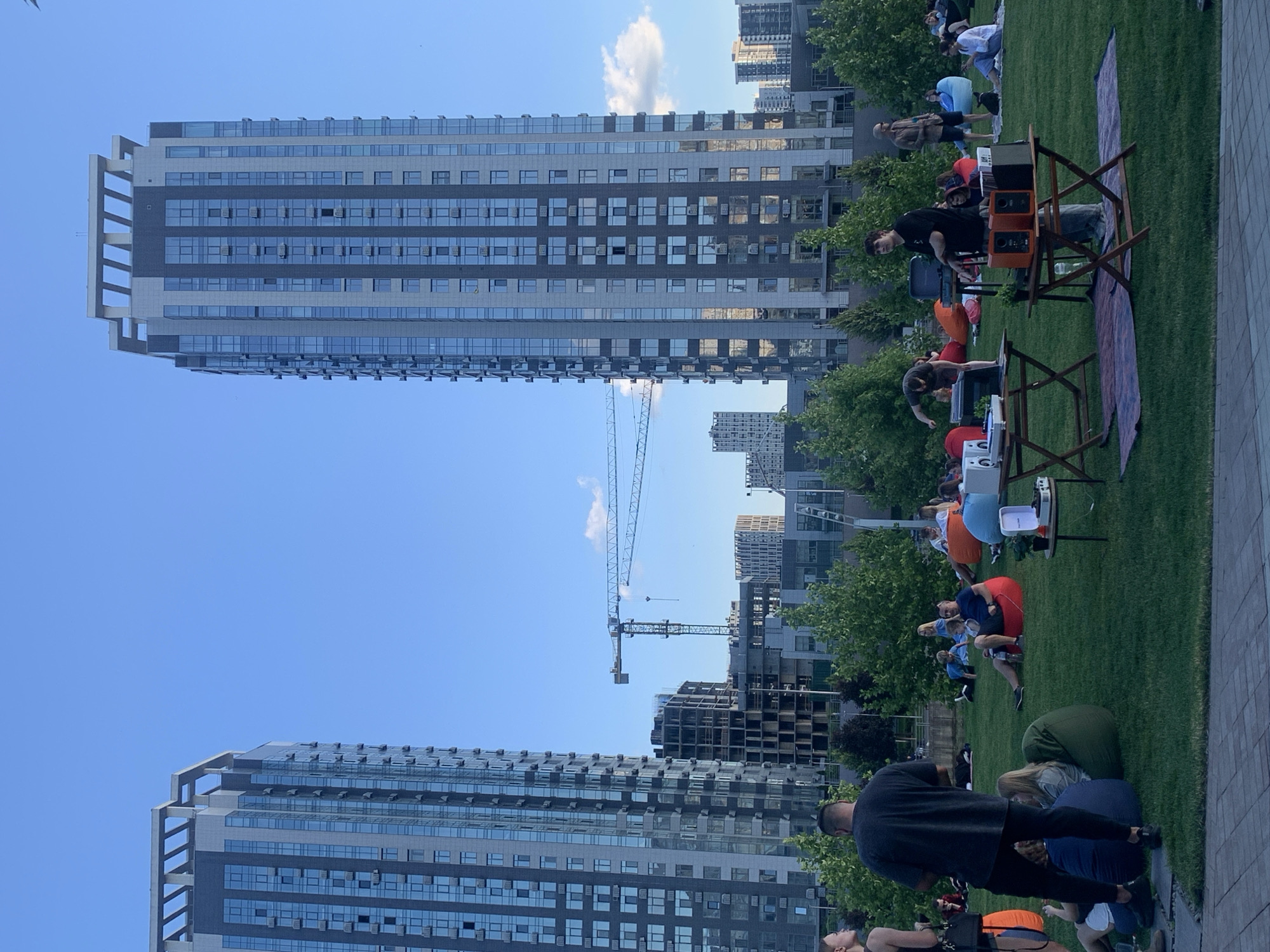 This is a wide shot of an outdoor event taking place in a park, in the city of Kyiv, Ukraine, with modern high-rise buildings in the background. In the foreground, a paved walkway leads to a large, well-maintained grassy area where approximately 30-40 people are gathered. Most of the attendees are seated on colorful beanbag chairs, or on blankets spread on the grass, facing a small setup in the middle of the lawn. Two wooden tables are positioned side-by-side, with electronic equipment, including what appears to be a DJ setup with turntables and speakers, placed on them. One person, a young male wearing a black t-shirt and jeans, stands by the tables, seemingly operating the equipment. Another person, a male wearing a black t-shirt, is visible in the mid-ground, also by the setup. The overall atmosphere suggests a relaxed gathering, possibly an outdoor movie screening or a music event. The backdrop features lush green trees, and beyond them, several contemporary apartment buildings, some of which are still under construction, indicated by visible cranes and scaffolding. The sky is clear and blue with a few wispy clouds.
