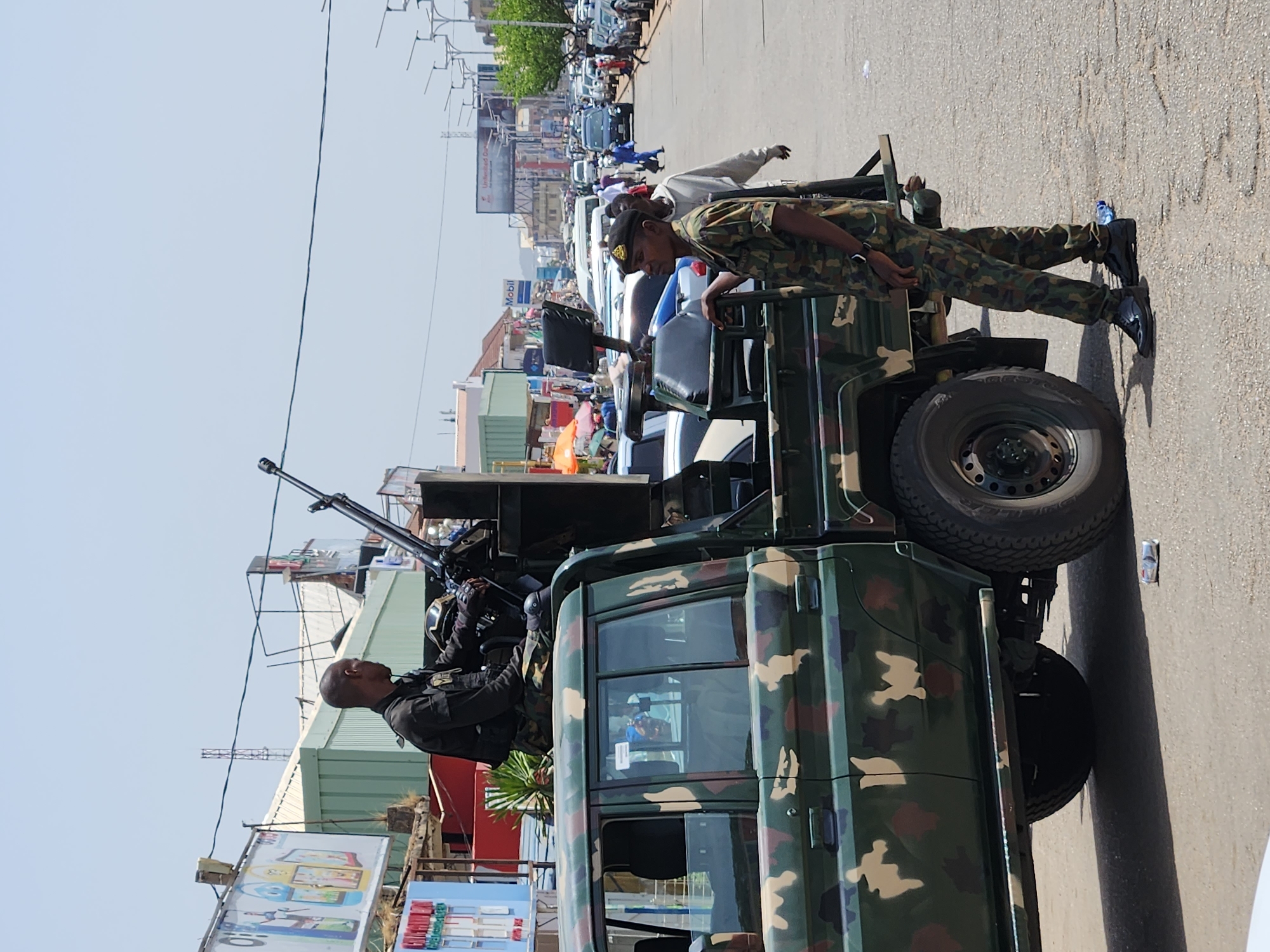 A daytime outdoor medium shot captures a military vehicle situated on a city street in Unknown, Unknown. The vehicle features a camouflage pattern, with two uniformed individuals. One individual is positioned on top of the vehicle operating a mounted weapon, while the other stands on the side, seemingly supervising or monitoring the surroundings. The street behind the vehicle is lined with various buildings, shops, and a dense array of vehicles and pedestrians, indicating a bustling urban environment. The sky is clear and a power line is visible above the street.
