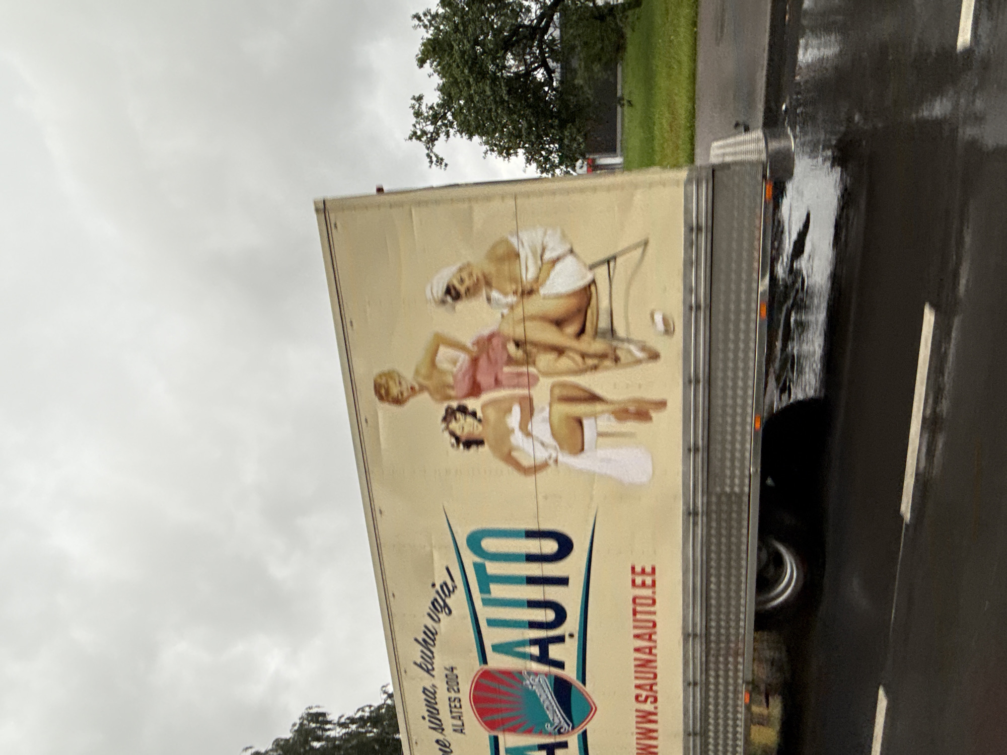The image captures a side view of a trailer truck parked on a wet, asphalt road, likely in Tallinn, Estonia, under an overcast sky. The truck's side is adorned with a large advertisement featuring stylized illustrations of three women in vintage pin-up style. Two women are depicted sitting, wrapped in towels, while another stands behind them, wearing a pink dress and a towel around her hair. The advertisement includes text in Estonian, with "SAUNA AUTO" prominently displayed in large teal and dark blue lettering, along with a red and teal shield logo. Below this, the website address "WWW.SAUNAAUTO.EE" is visible in red text. The overall environment suggests a somewhat gloomy or rainy day, as indicated by the wet road and the gray sky. Trees and grass are visible on the right side of the image, bordering the road.